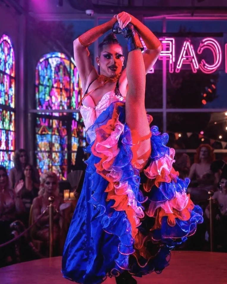 Une danseuse porte une robe colorée avec des épaules dénudées et de longues gants en dentelle. Elle réalise une pose avec une jambe levée en haut de la tête dans un décor de théâtre avec vitraux colorés en arrière-plan et un panneau néon rose "CATH".