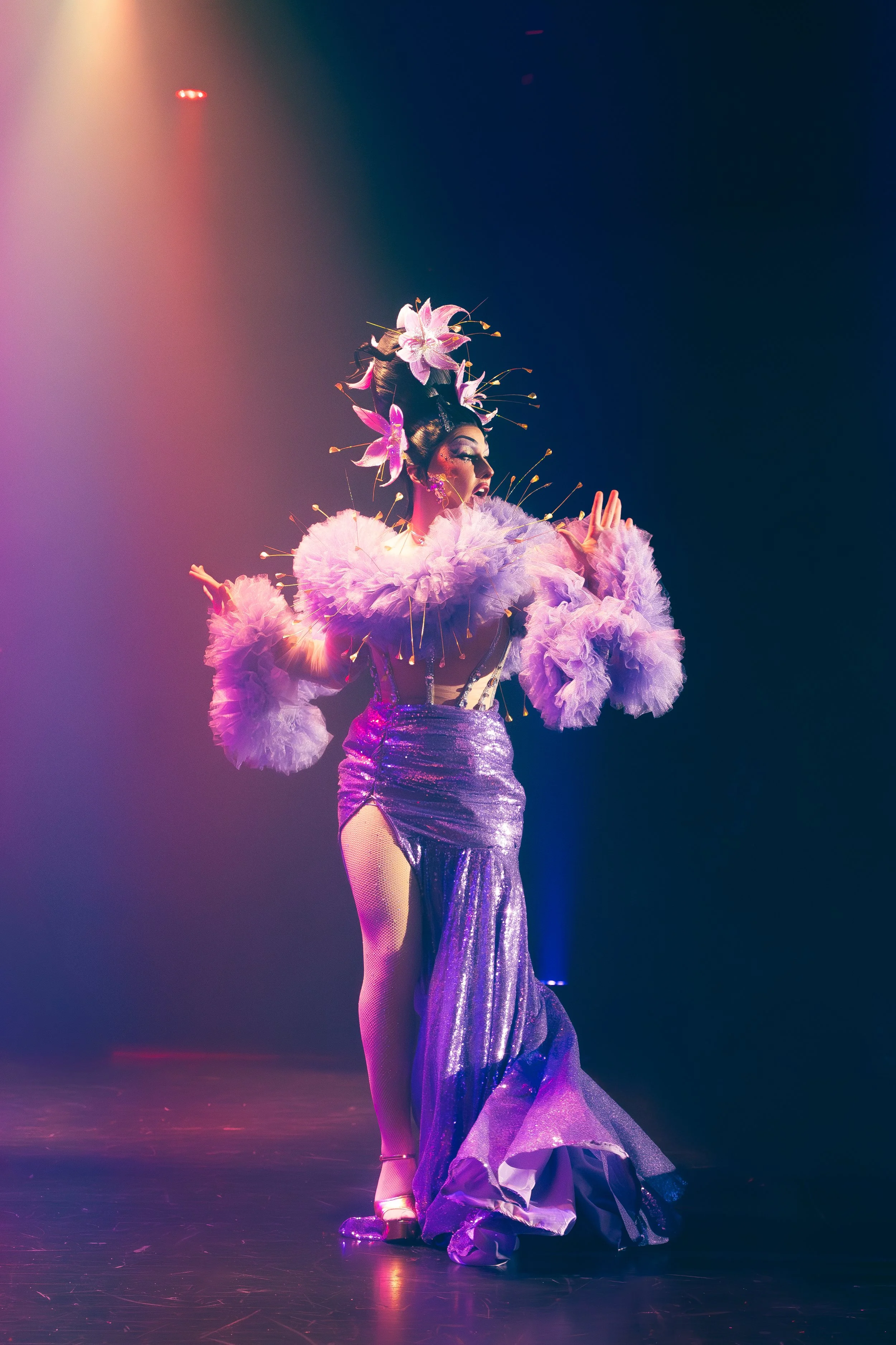 Une performeuse en costume rose et violet, avec des fleurs sur la tête, se tient sur scène dans un éclairage coloré.