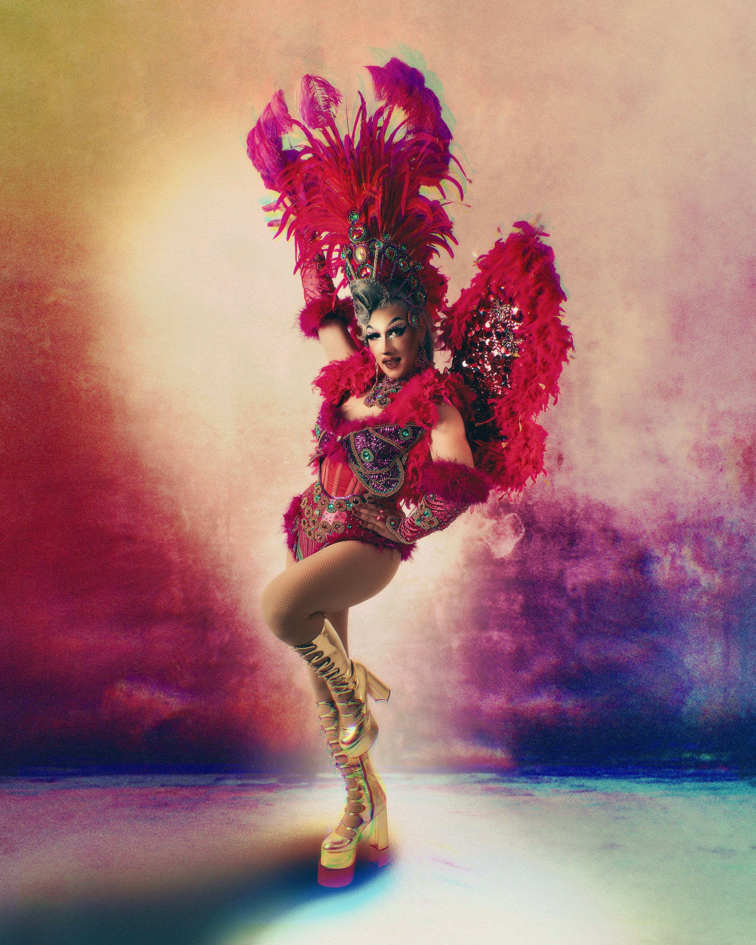 Kitsch, drag queen, en costume de spectacle avec un grand chapeau orné de plumes colorées, portant une robe embellie de bijoux, en pose dynamique avec un fond multicolore abstrait.