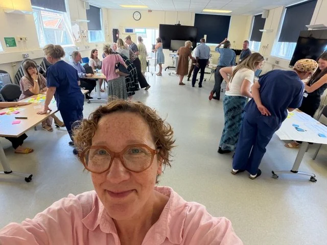 Jessie Cunnett, a woman with curly hair and glasses, takes a selfie in a bright room filled with people engaged in activities and discussions.