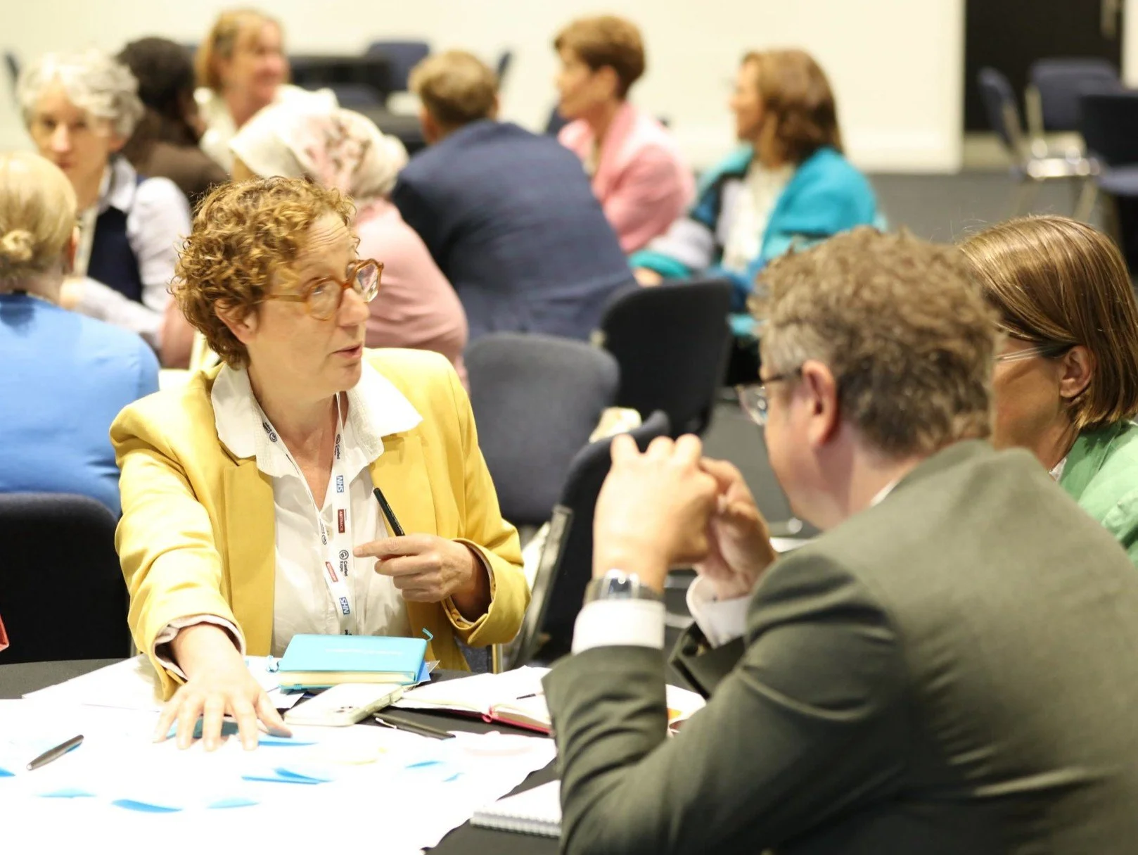Multiple people engaged in discussions at a workshop, with Jessie explaining the documents in front of her.