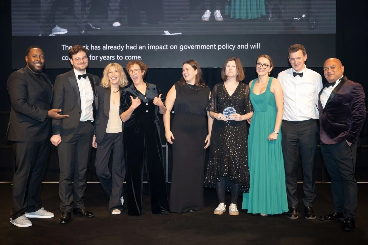 Group of nine people dressed formally accepting an award at a ceremony, standing on a stage with a large screen behind them. Jessie Cunnett gives a thumbs up.
