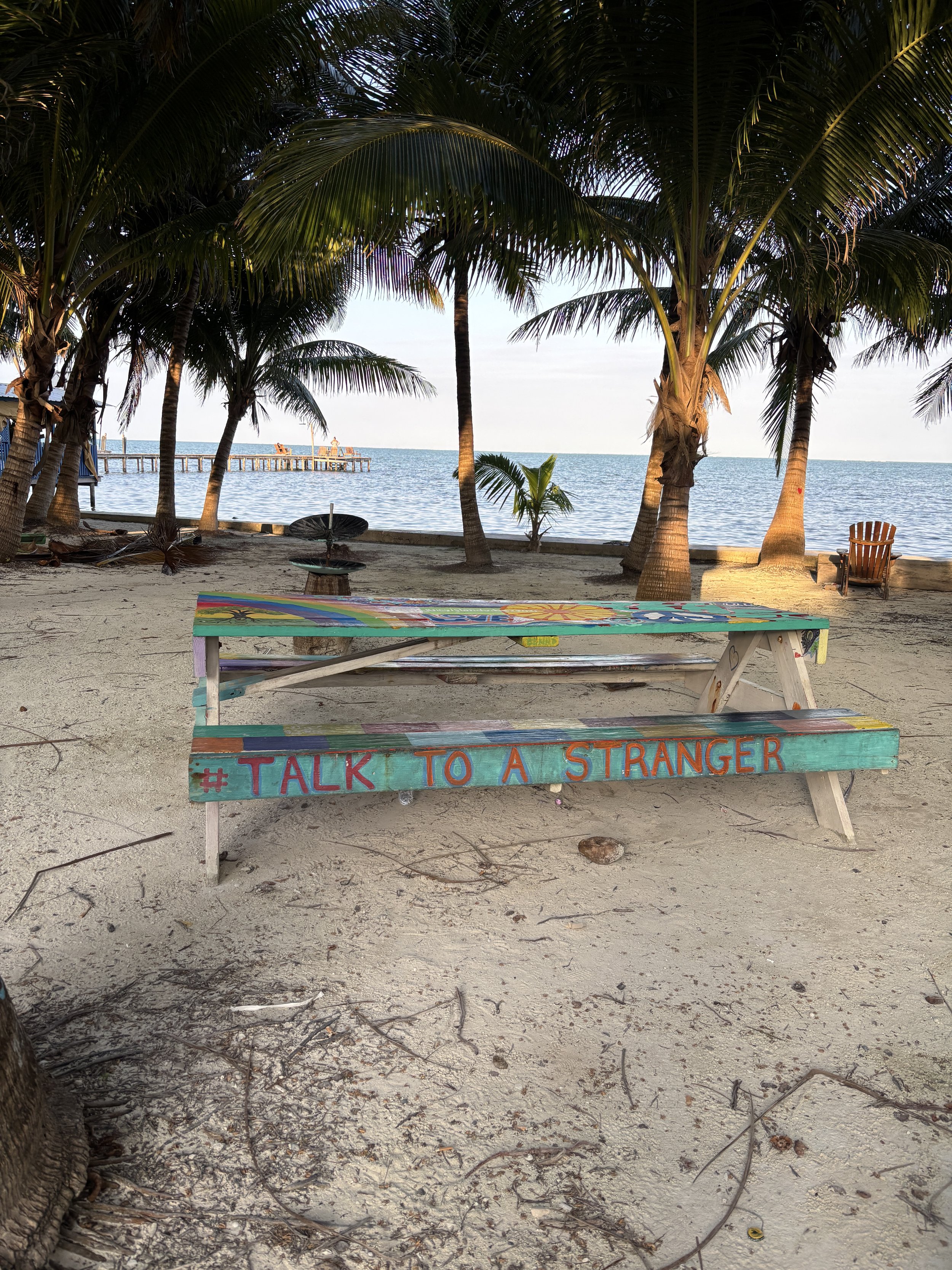 Colorful sign on a picnic table on a sandy beach with palm trees and a pier in the background.