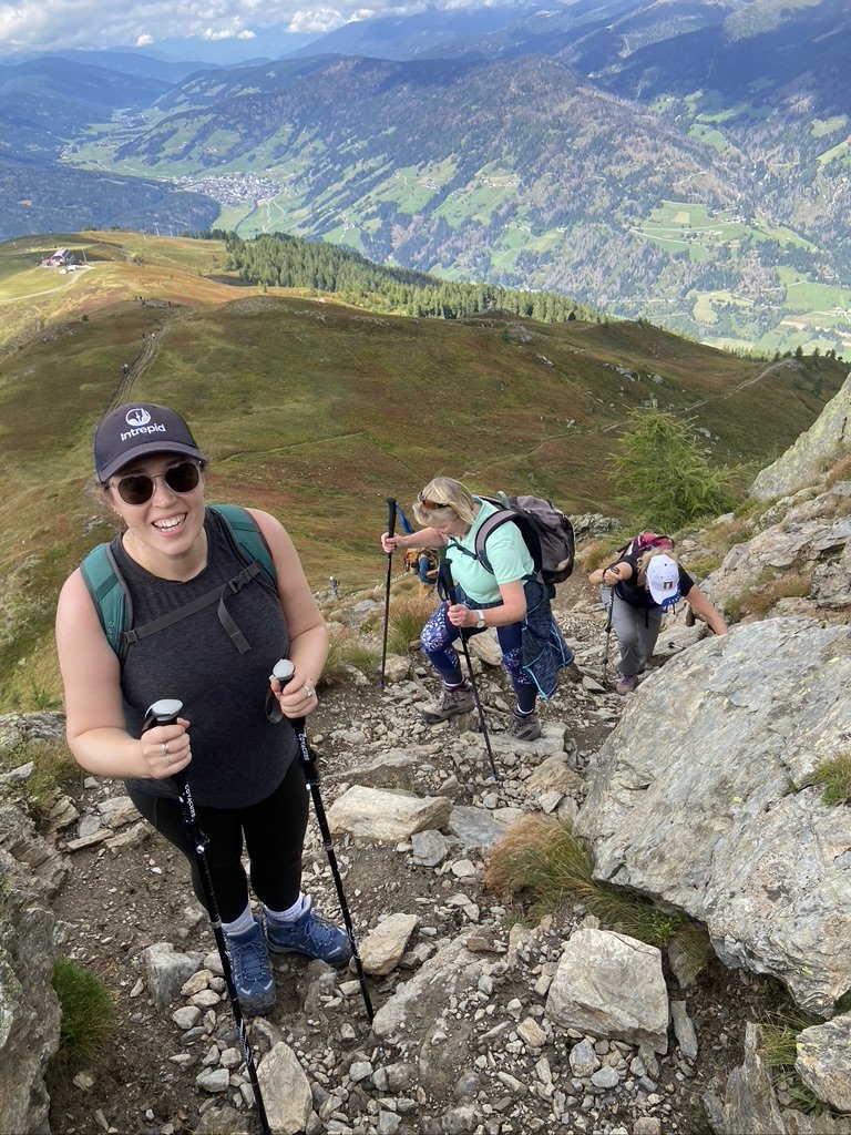 Smiling woman (Morwenna) hiking on rocky trail in the Dolomites with mountain and valley view in background, accompanied by three other hikers ascending behind her.