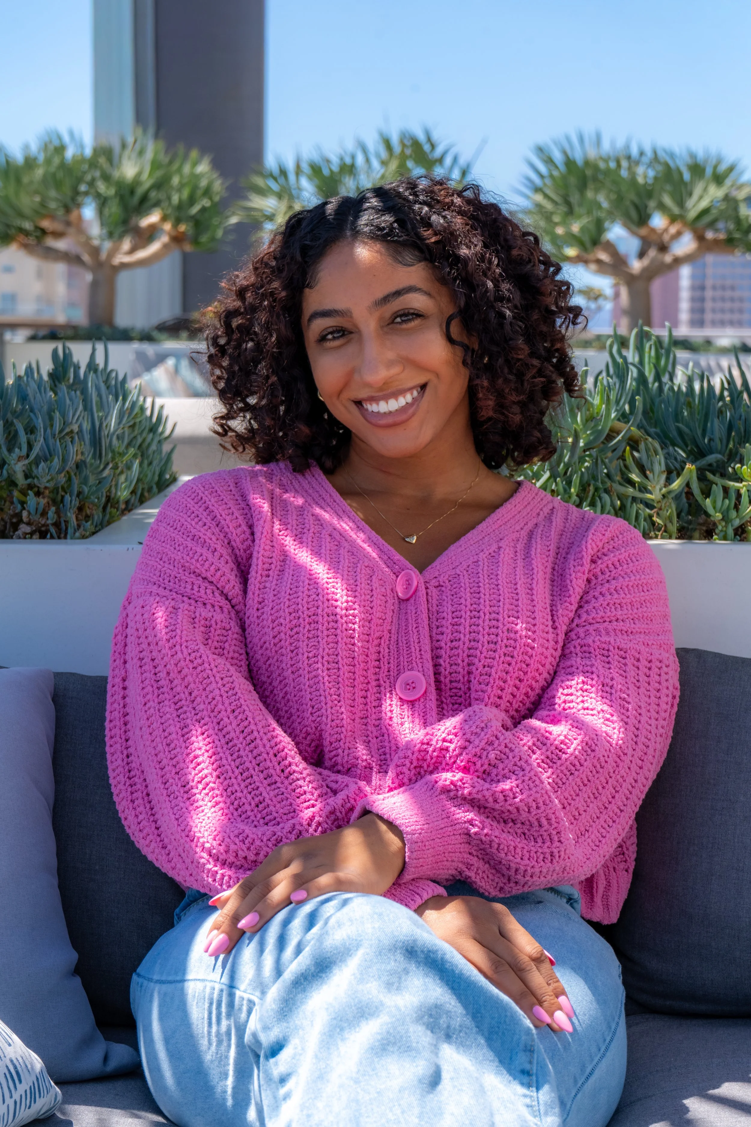 A woman with curly dark hair wearing a pink knitted sweater, sitting outdoors on a grey cushion with her hands crossed and smiling toward the camera, surrounded by green plants and trees against a cityscape background on a sunny day.