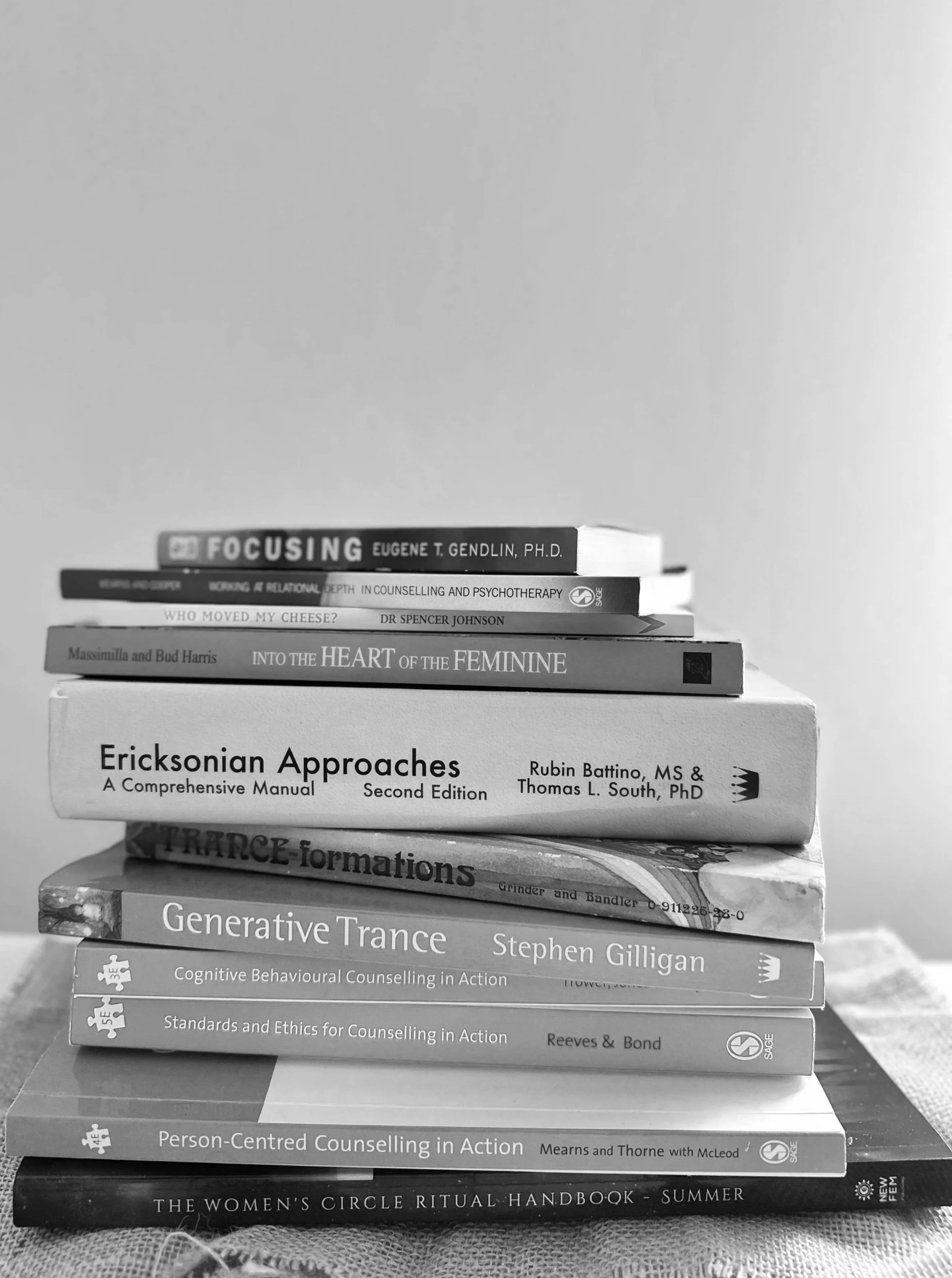 Stack of books on counseling, therapy, and personal development topics with the titles: 'Focusing' by Eugene T. Gendlin, 'Wrong at Relational Depth' by Unknown, 'Who Moved My Cheese' by Dr. Spencer Johnson, 'Into the Heart of the Feminine' by Massimilla and Bud Harris, 'Ericksonian Approaches' by Rubin Battino and Thomas L. South, 'Trance-formations' by Grinder and Bandler, 'Generative Trance' by Stephen Gilligan, 'Person-Centred Counselling in Action' by Mearns and Thorne, and 'The Women's Circle Ritual Handbook-Summer'.
