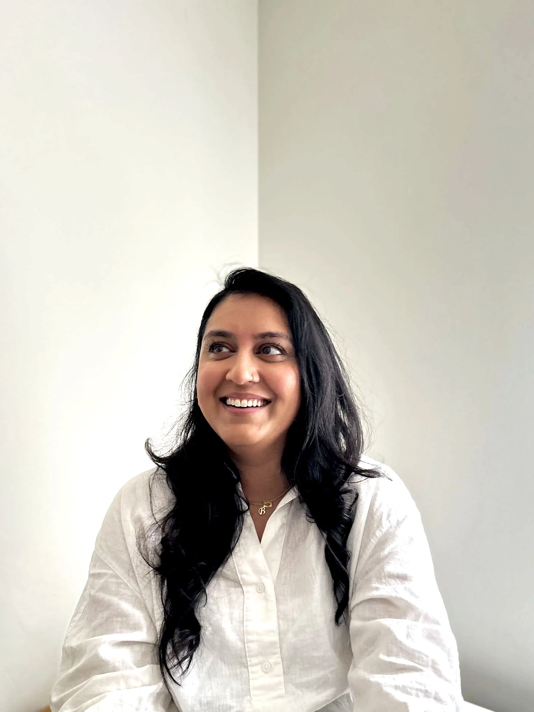 A woman with long dark hair and a light skin tone smiling, wearing a white button-up shirt and a gold necklace with a B pendant, sitting against a plain light-colored wall.