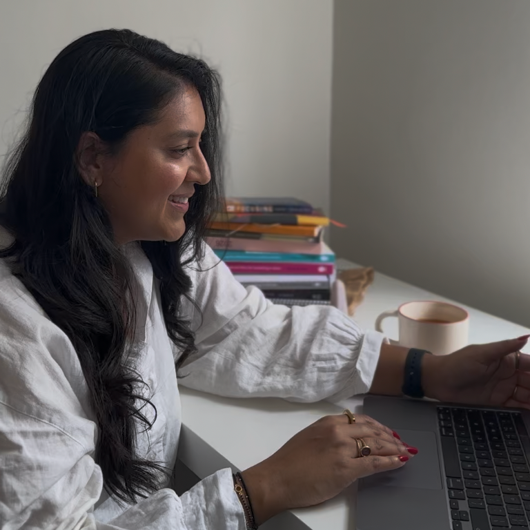 A woman with long dark hair smiling while working on a laptop at a desk with a stack of books and a mug nearby.