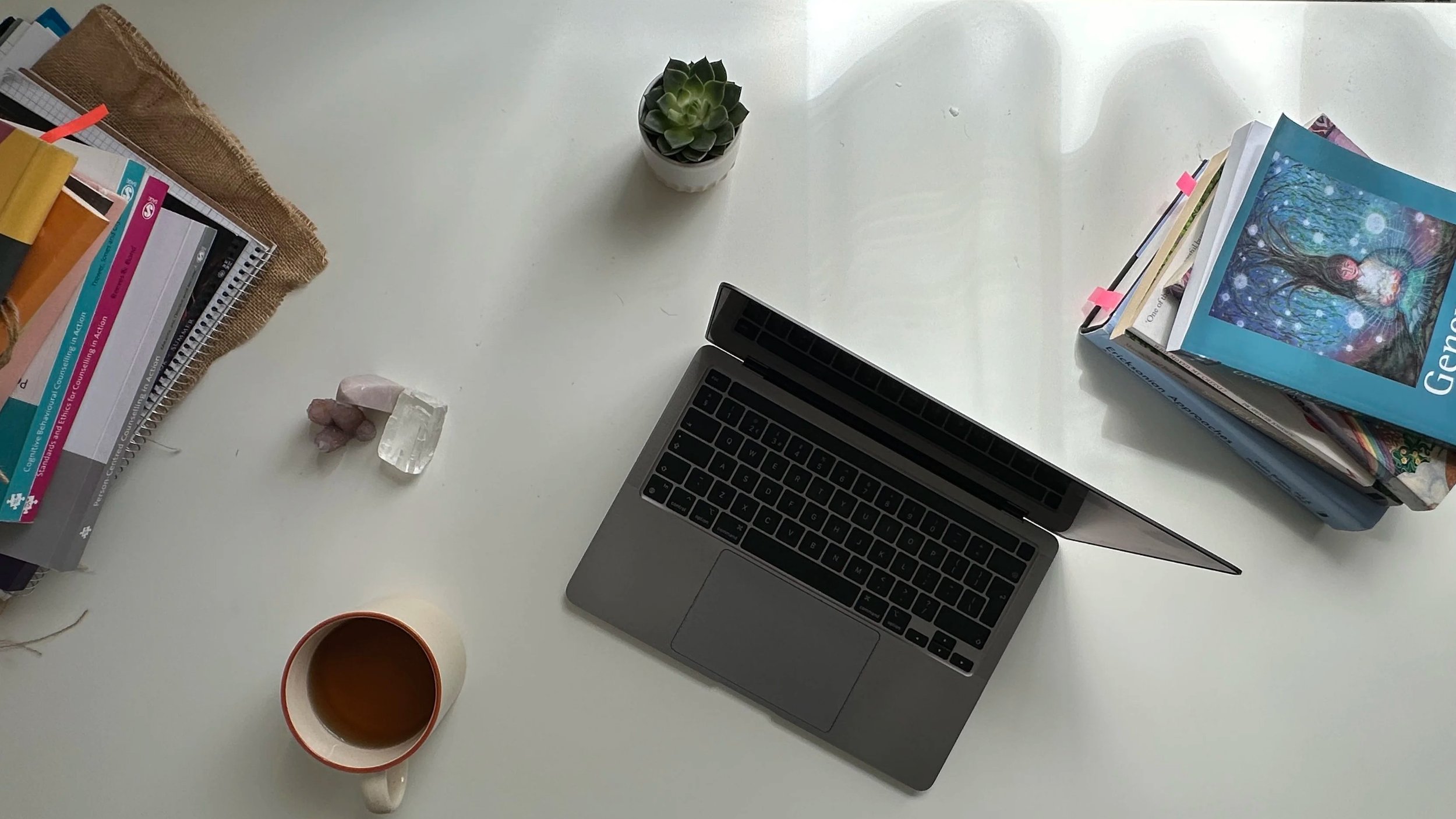 Desk with open laptop, books, potted succulent, stack of notebooks, crystals, and a cup of tea.