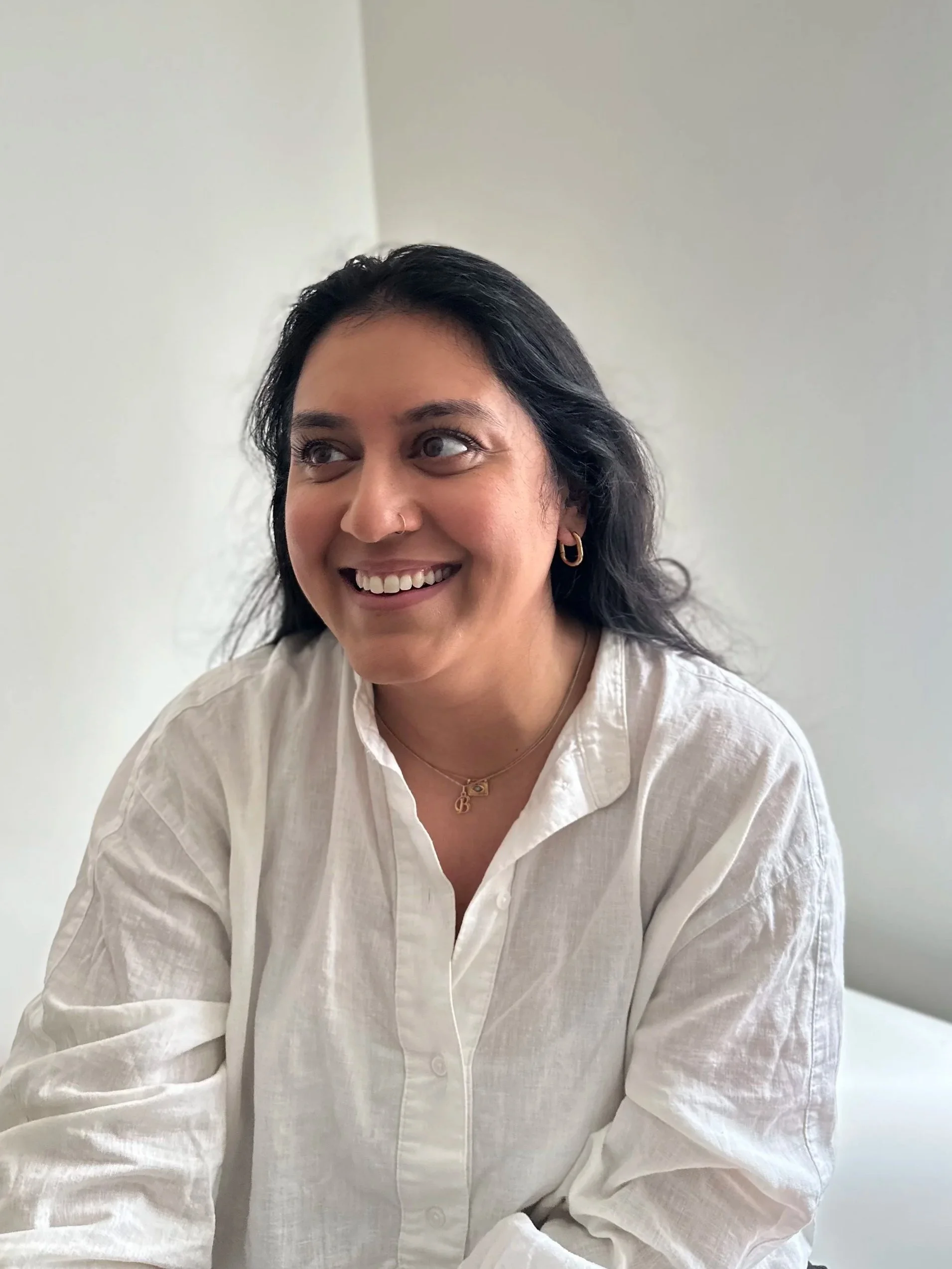 A woman with dark hair, smiling, wearing a gold hoop earring, a small nose ring, a gold necklace with a pendant, and a white shirt, sitting indoors against a plain white wall.