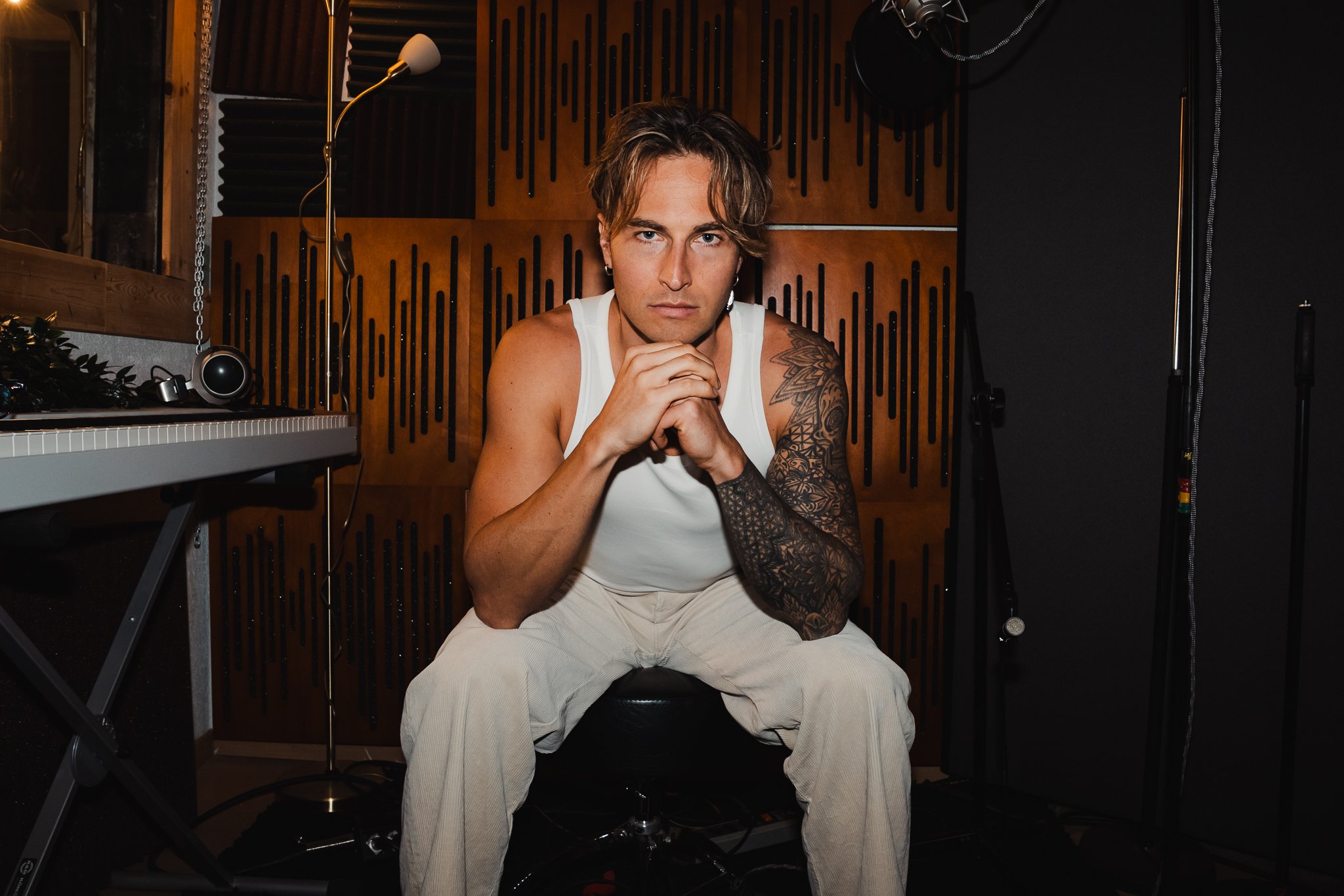 A man with tattoos and long hair sitting in a recording studio, wearing a white tank top and beige pants, with his hands clasped under his chin, looking directly at the camera.