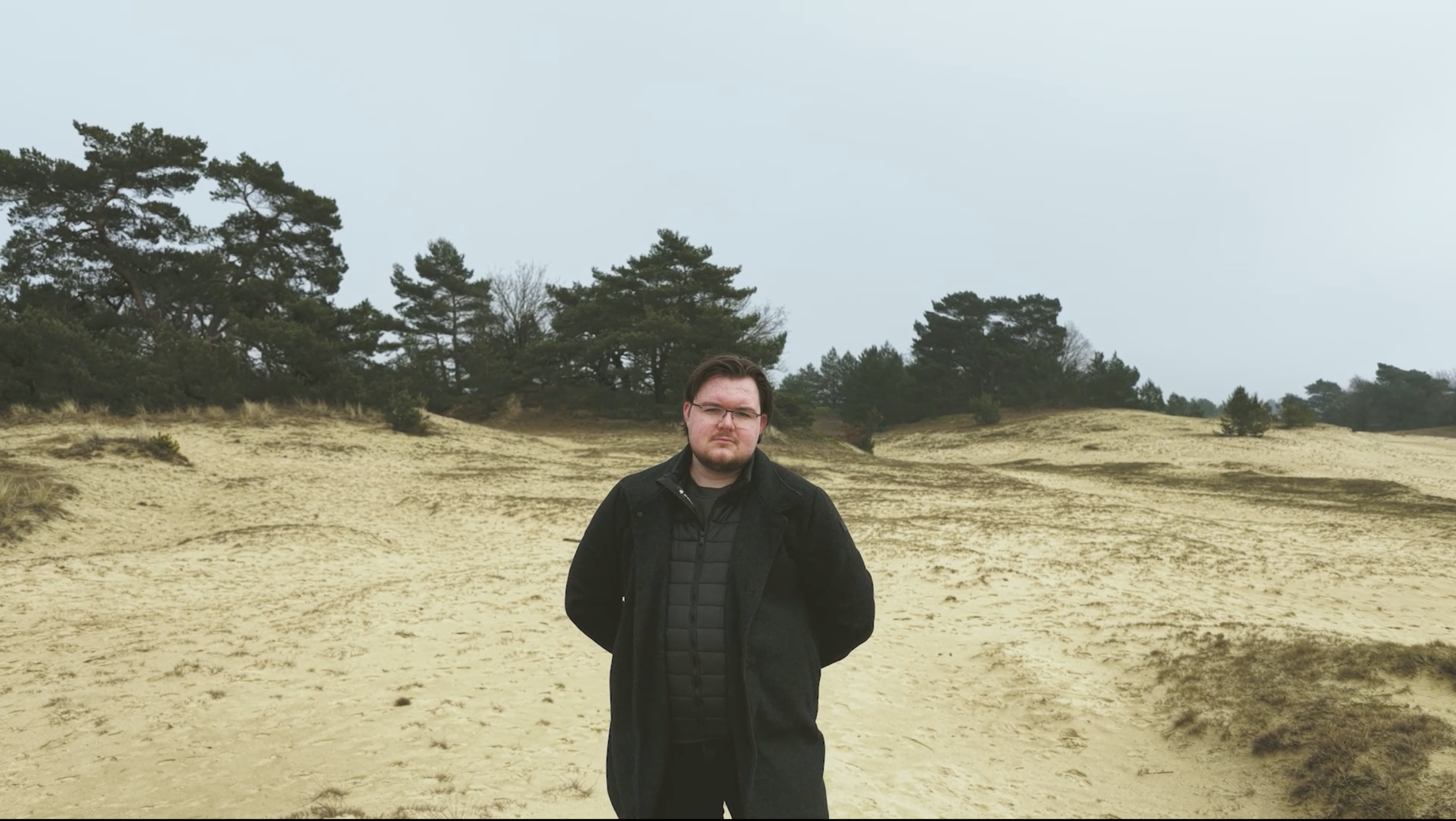 Man staat in een zandlandschap met dennenbomen op de achtergrond, draagt een zwarte jas en bril, kijkt naar de camera.