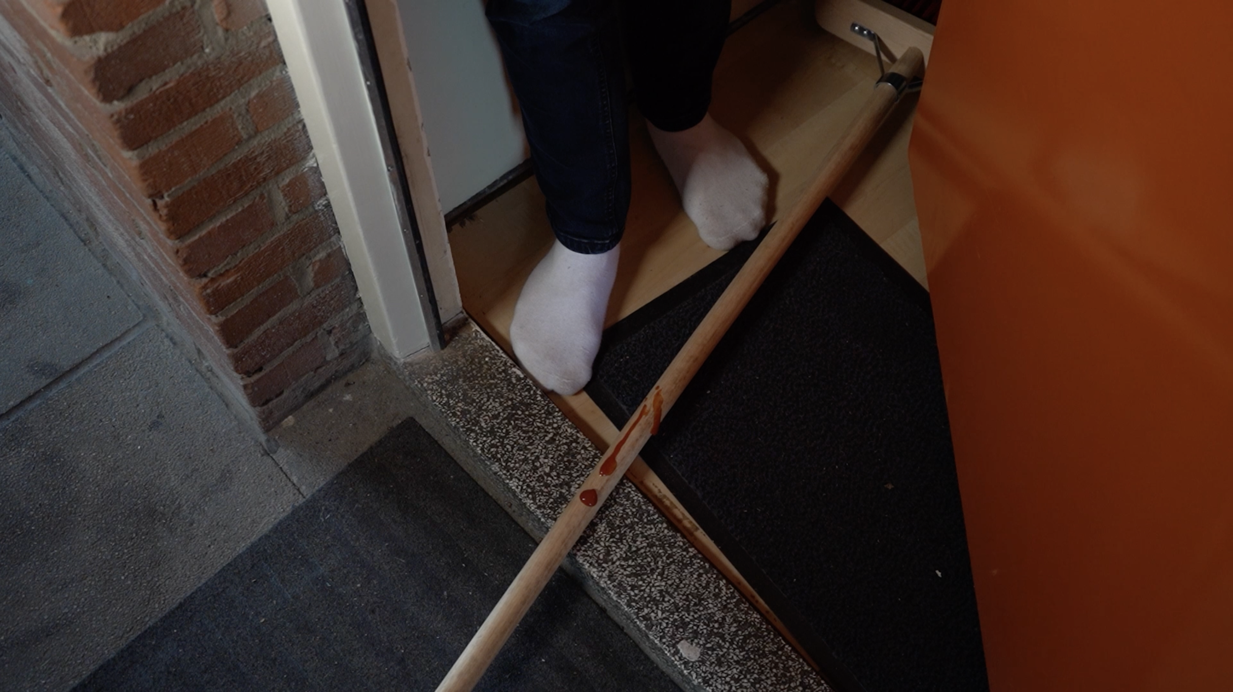 Persoon zit op een houten bankje, draagt witte sokken en een donkere broek, naast een muur met bakstenen en een deur. Op de vloer ligt een zwarte mat.