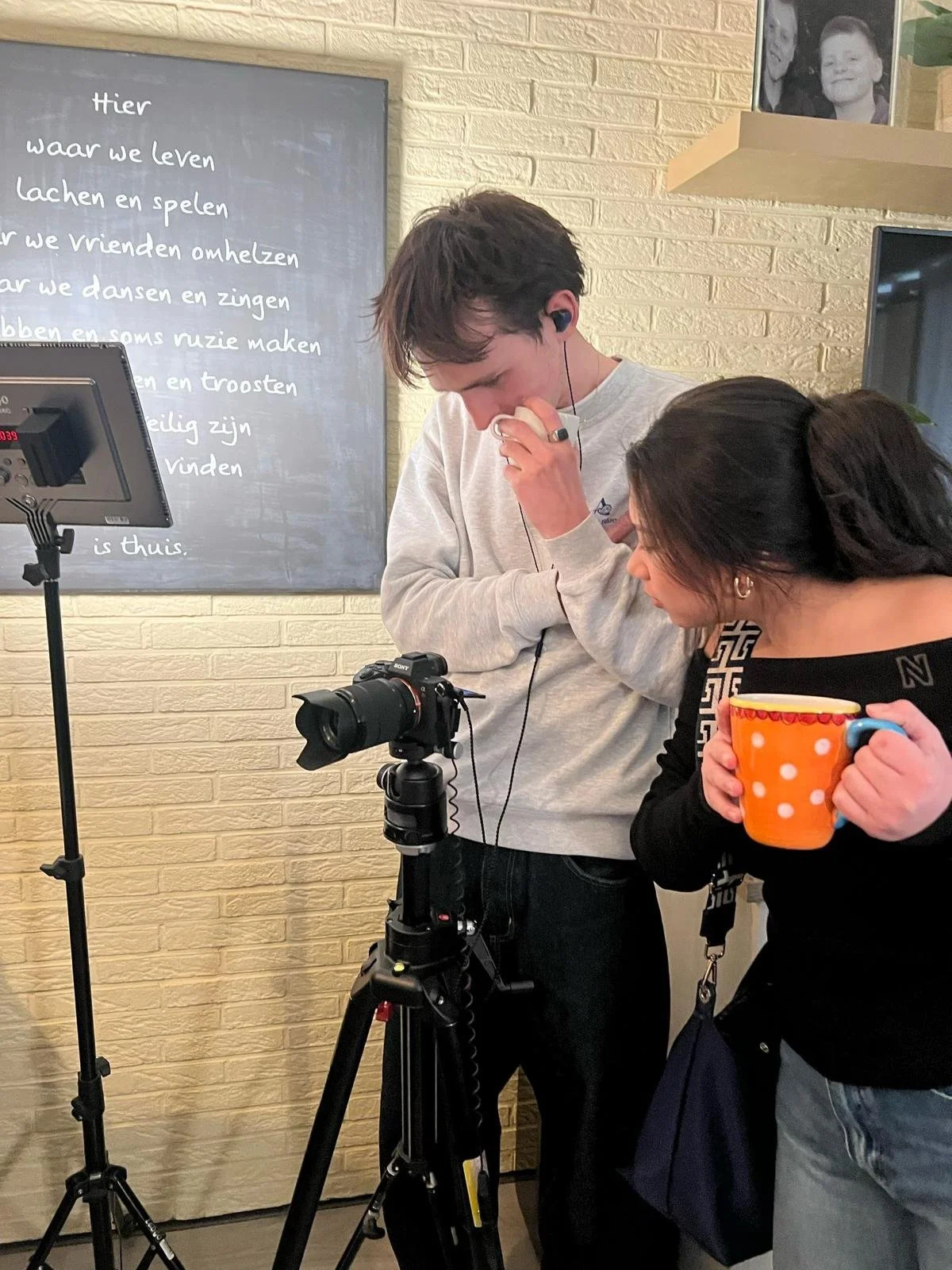 Twee mensen kijken naar een camera die op een statief staat, terwijl een vrouw een grote oranje mok vasthoudt. Op de achtergrond een muur met een zwartbord met witte tekst, een foto op een plank, en een televisie.