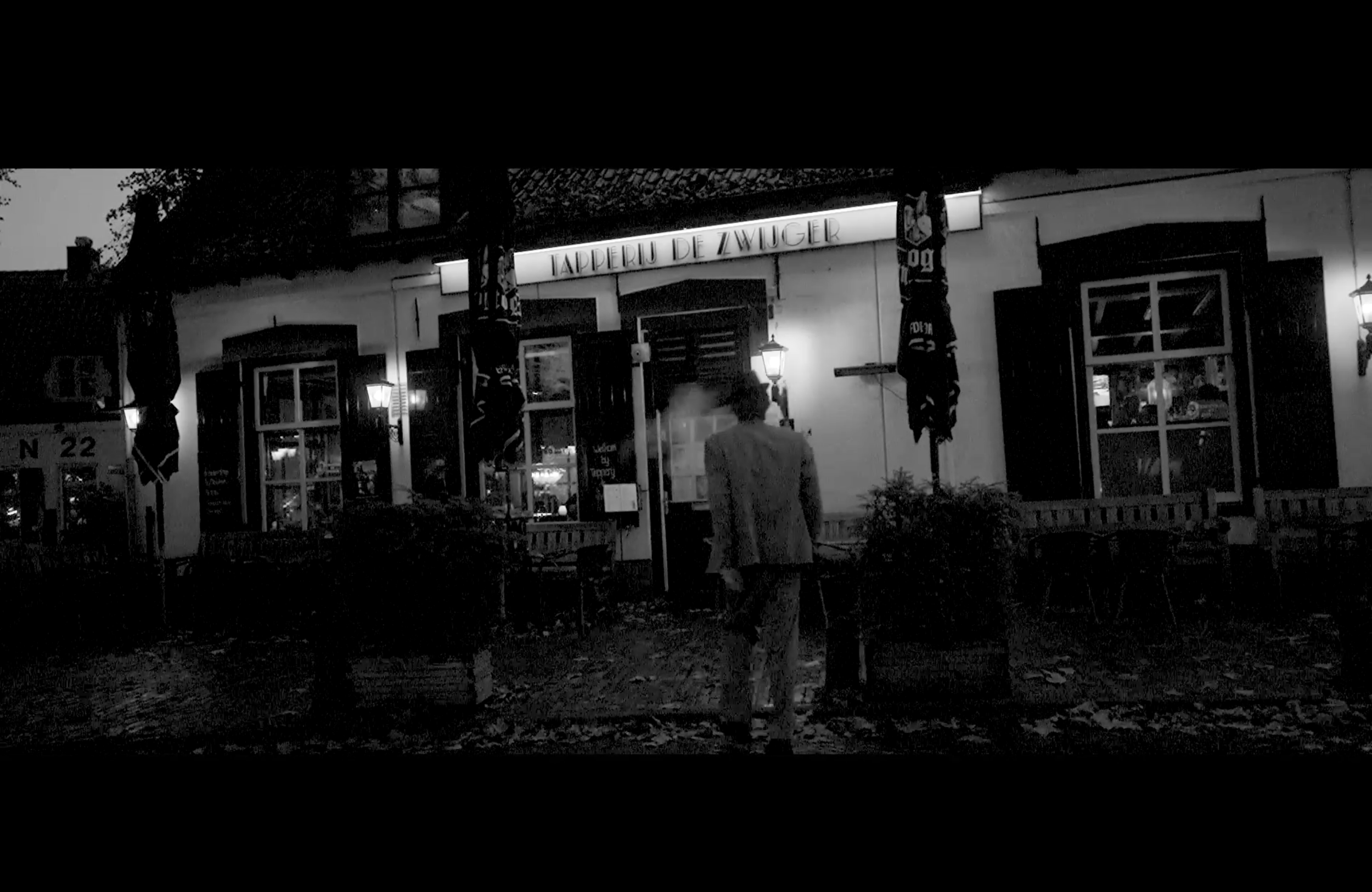 Een zwart-wit foto van een restaurant met de naam 'Tapperij De Zwalger', buitenverlichting en mensen die naar binnen gaan.