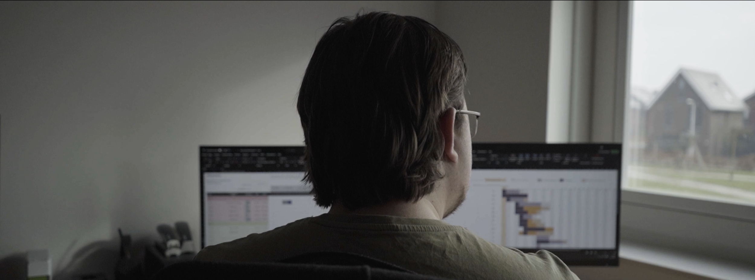 Man zit aan een bureau, kijkt naar een computerscherm met grafieken en datavisualisaties, voor een raam met huizen op de achtergrond.