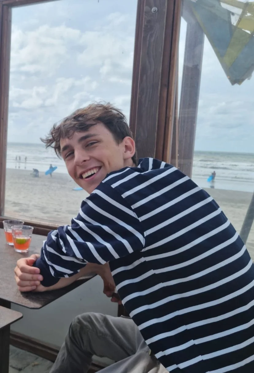 Een jongen die glimlacht aan de kust terwijl hij binnen zit, met water en parasols op het strand op de achtergrond.