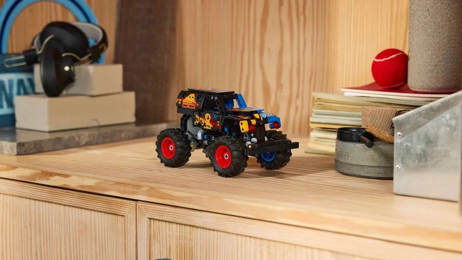 LEGO Monster Jam Grave Digger Fire and Ice truck in black and blue with flame graphics and red wheels, sitting on a wooden cabinet beside headphones and books