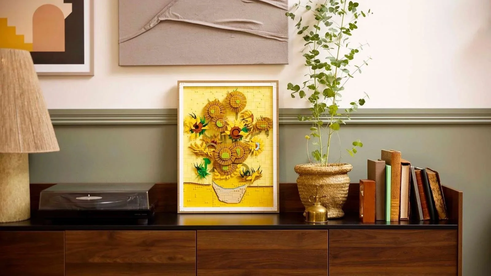 LEGO Vincent van Gogh Sunflowers mosaic in a wood frame, displayed on a dark wood sideboard beside a eucalyptus plant and stacked books