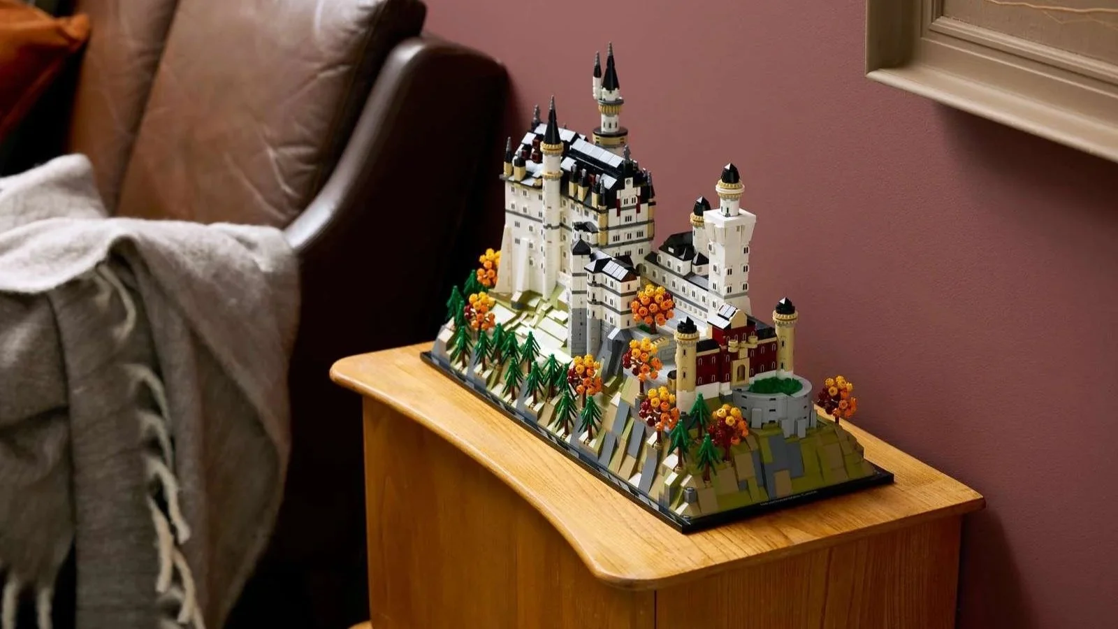 LEGO Neuschwanstein Castle model surrounded by autumn trees, displayed on a wooden nightstand beside a leather armchair with a gray throw