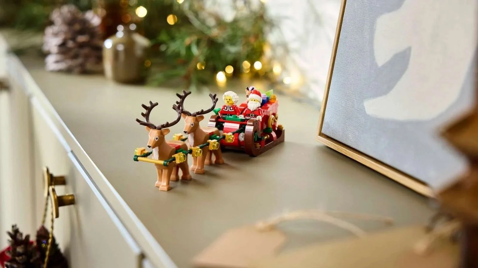 LEGO Santa sleigh set with two reindeer, Santa and Mrs. Claus on a sideboard with warm string lights behind