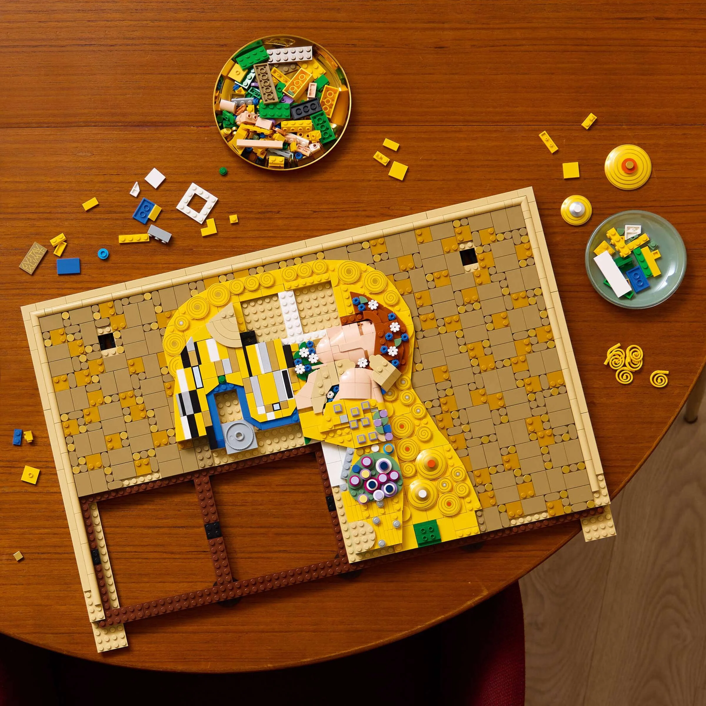 A LEGO Art build of Gustav Klimt's The Kiss taking shape on a round wood table. The couple's embracing faces and golden mosaic pattern are already formed, with loose pieces sorted into small dishes nearby.