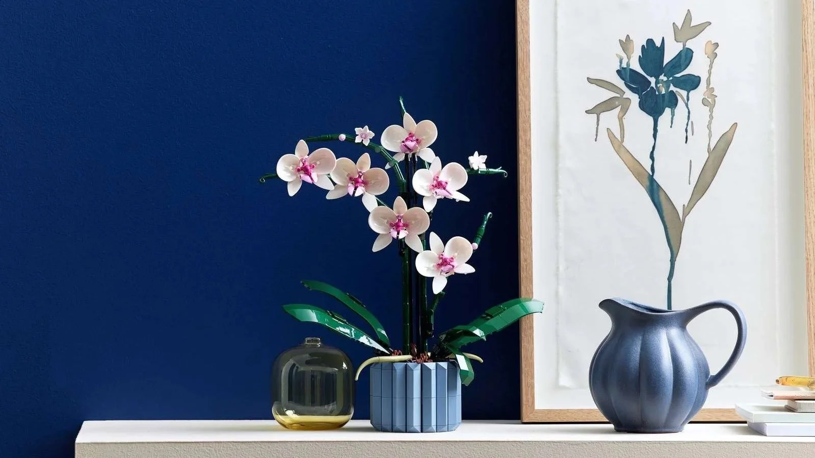LEGO Orchid set with white and pink blooms against a navy wall beside a framed botanical print