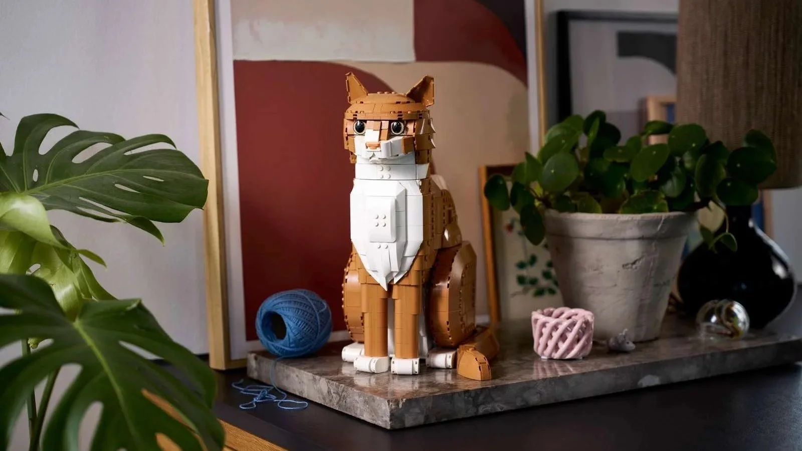 LEGO Orange Cat set, a seated ginger and white cat, displayed on a marble tray next to a potted plant and a ball of blue yarn