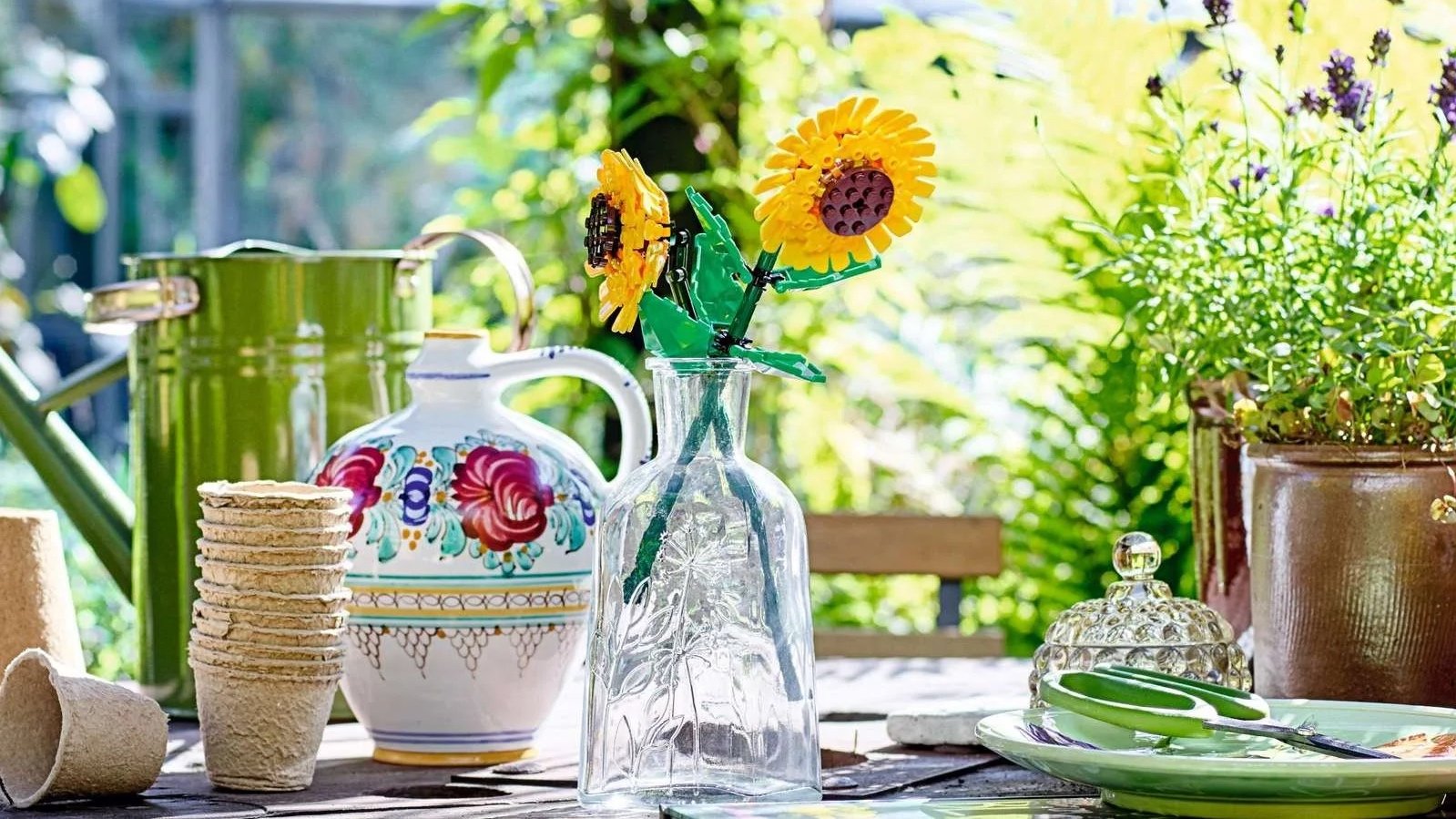 Two LEGO sunflowers in a clear glass vase on a garden table with a watering can and lavender