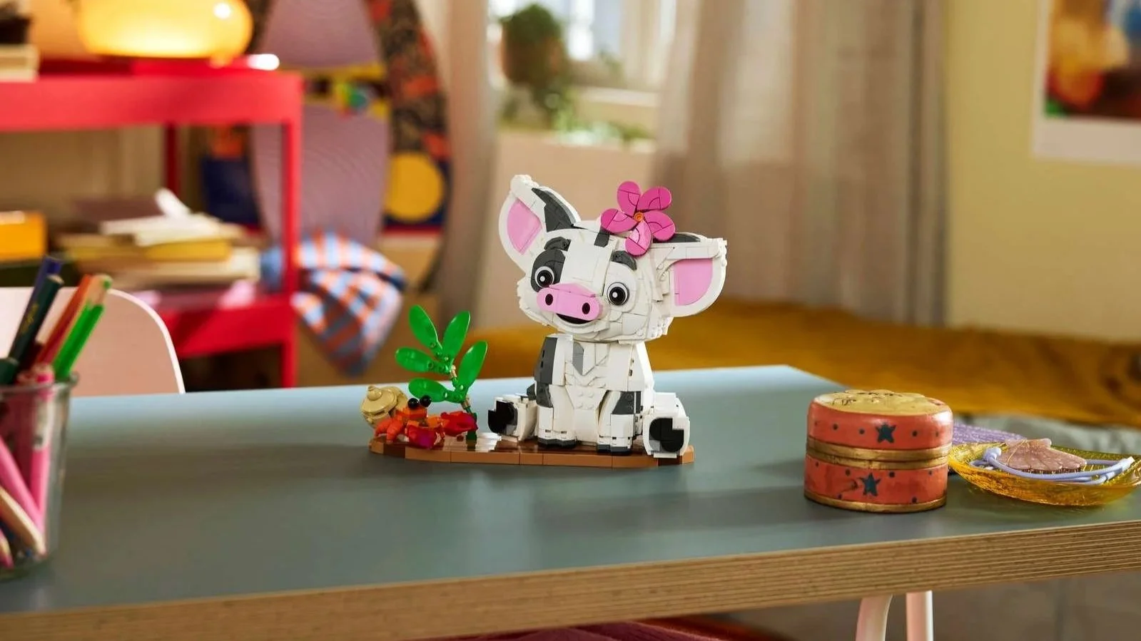 LEGO Pua set, a white pig with pink ears and a pink flower on its head, sitting next to a small crab and green leaves on a desk
