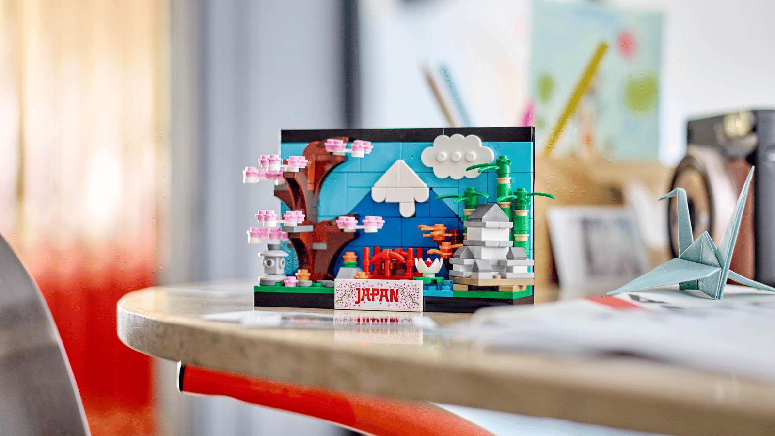 LEGO Japan postcard set with Mount Fuji, cherry blossoms, a pagoda and a red bridge, displayed on a desk next to an origami crane
