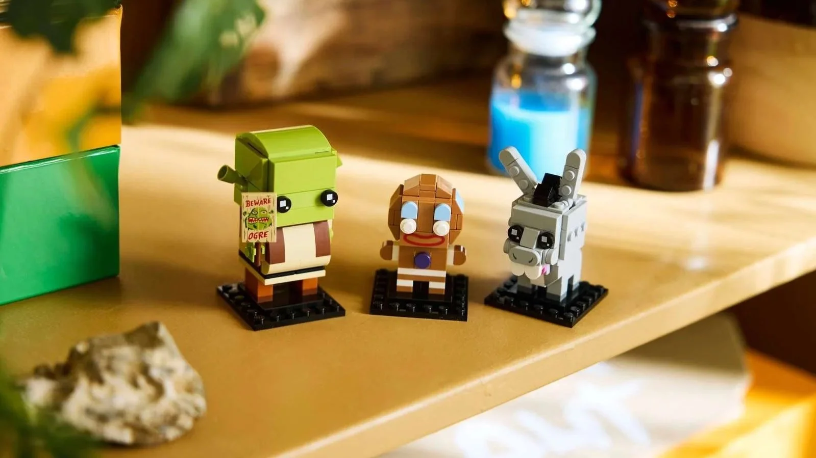 LEGO Shrek, Gingy and Donkey figures on black bases on a wooden shelf, Shrek with a Beware Ogre sign