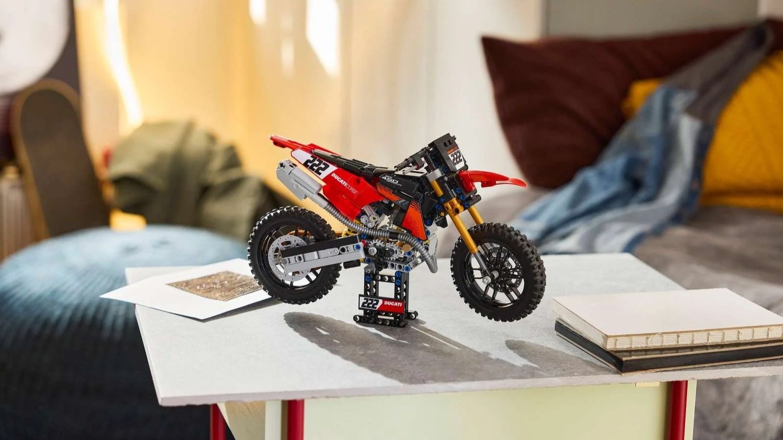 Red and white LEGO Ducati Desmo450 MX Factory dirt bike with number 222 on a display stand, sitting on a small table in a cozy bedroom
