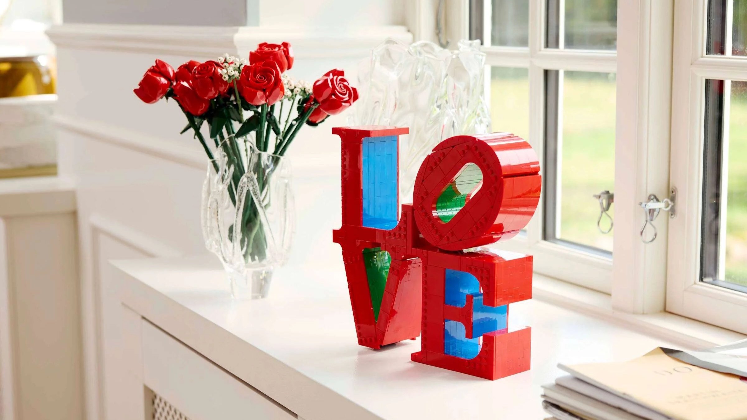 Red LEGO LOVE sculpture beside a vase of LEGO roses on a white counter by a bright window