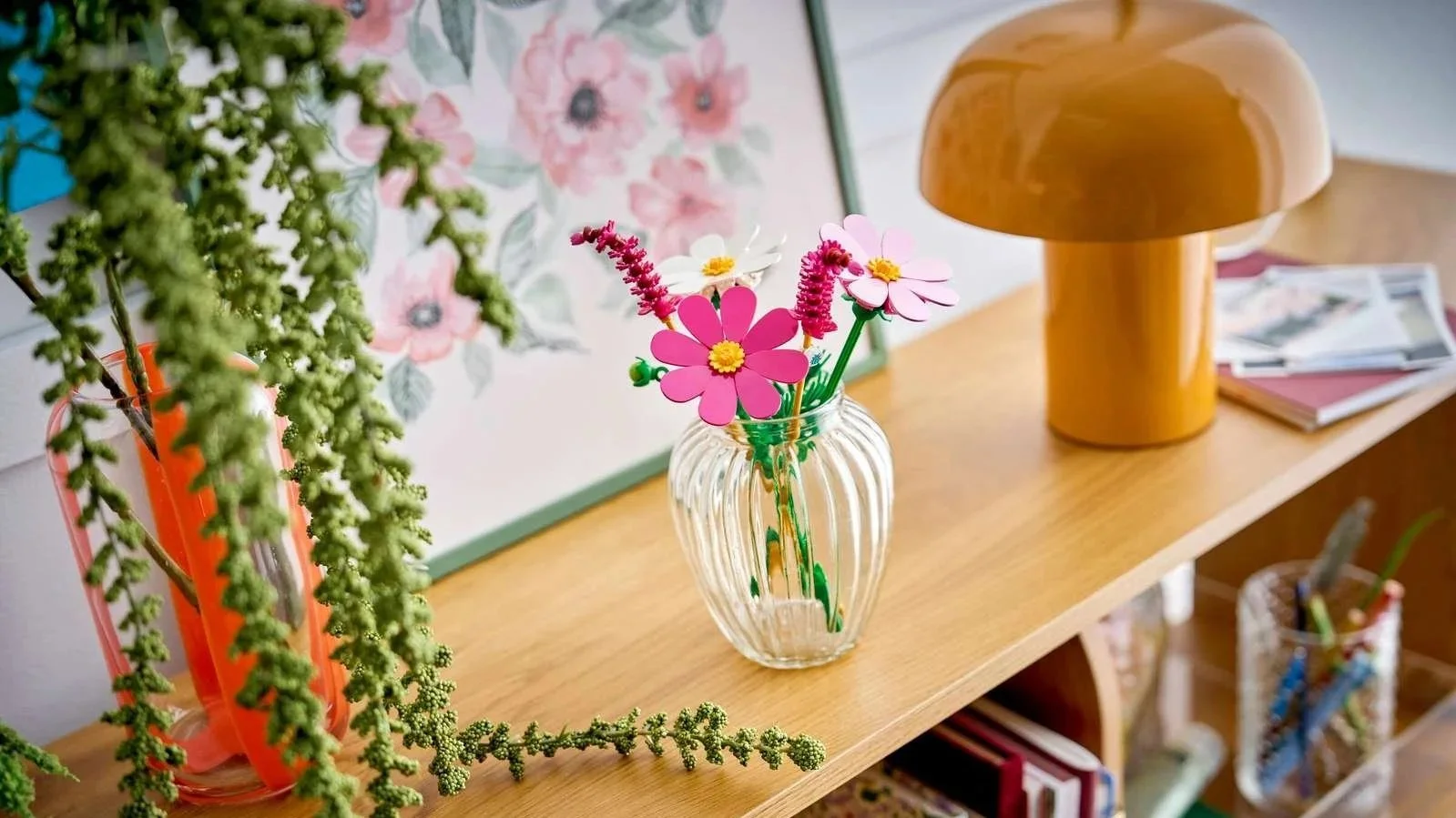LEGO Cosmos Flowers set in a ribbed glass vase on a wooden bookshelf beside a mustard mushroom lamp