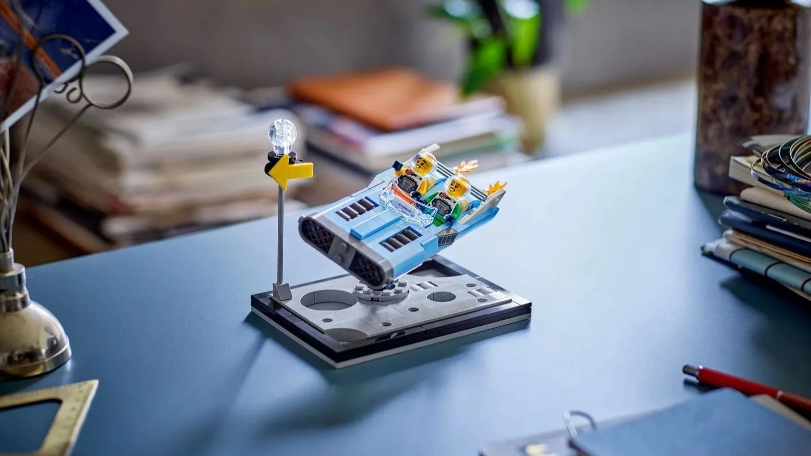 LEGO astronaut minifigures in a small blue and gray spacecraft, displayed on a desk with books, a plant, and a lamp