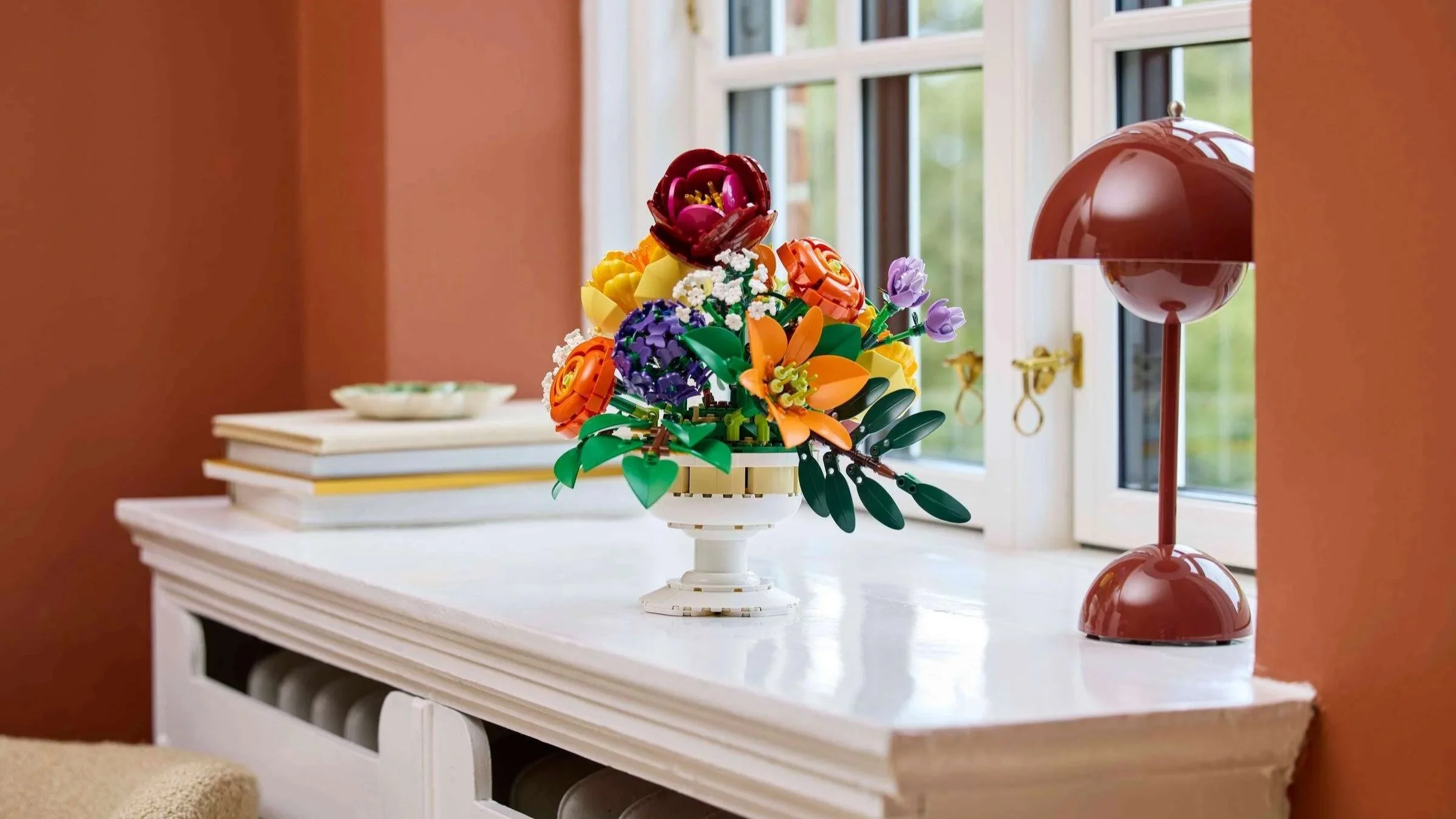 LEGO Flower Arrangement set in a white pedestal vase on a shelf by a window, styled next to stacked books and a red dome table lamp