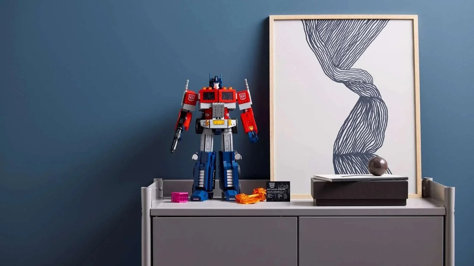 LEGO Optimus Prime set posed on a gray cabinet against a deep blue wall beside framed line art