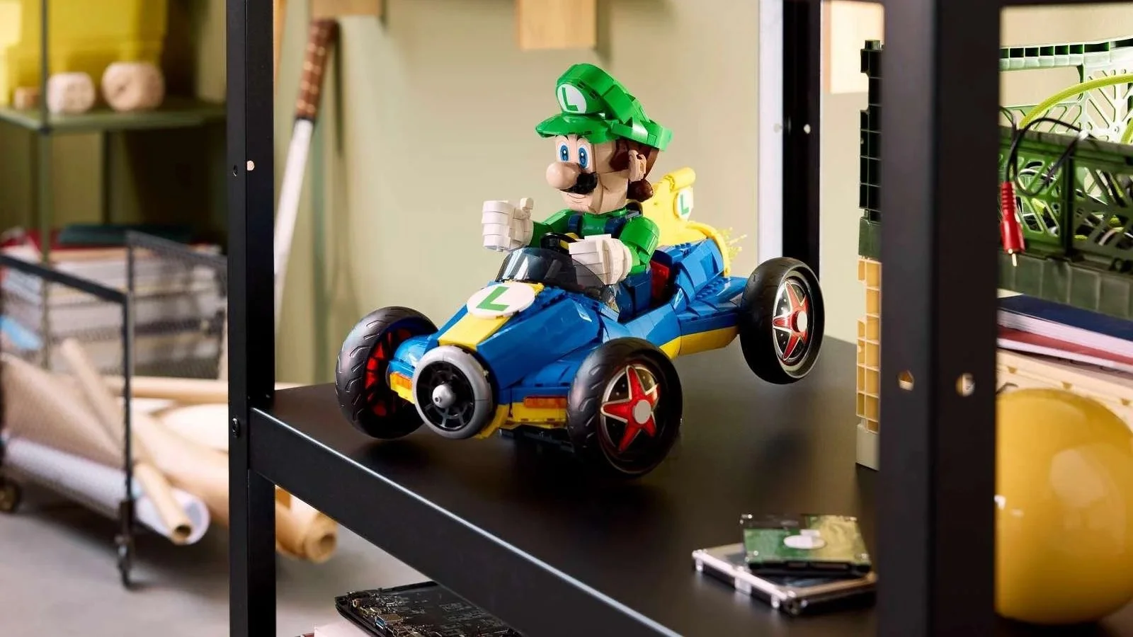 A LEGO Luigi figure drives his Mach 8 kart on a dark metal shelf in a workshop-style space, with tools and rolled paper nearby