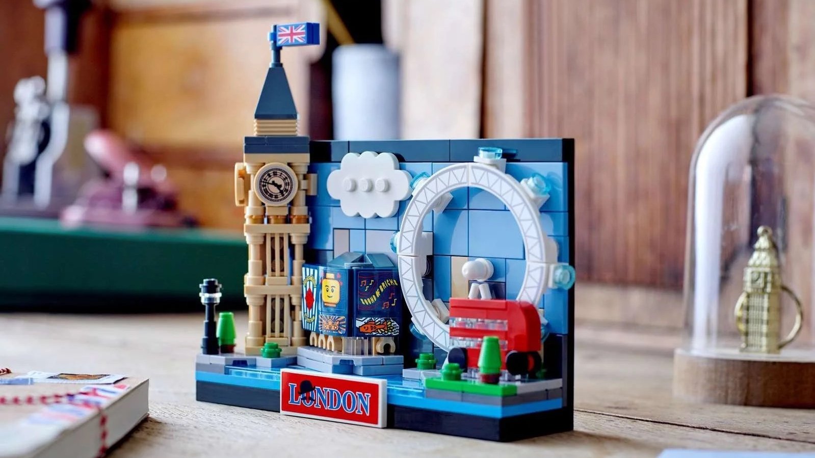 London Postcard LEGO set on a wooden desk with Big Ben, the London Eye, a red double-decker bus and a red London sign beside a notebook and pencil