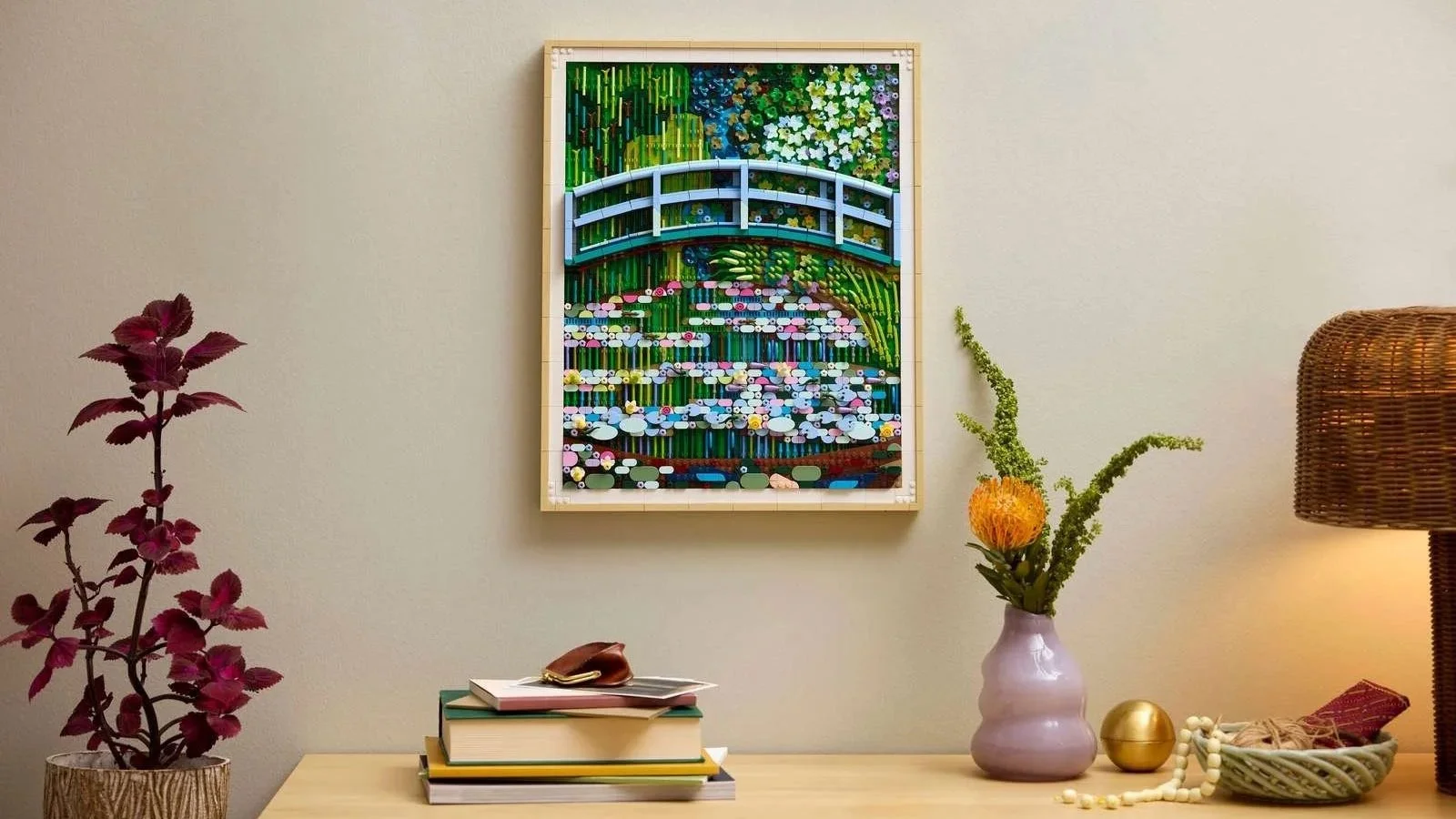 LEGO Claude Monet Bridge over a Pond of Water Lilies mosaic in a wood frame, hung above a table with books and a vase of flowers
