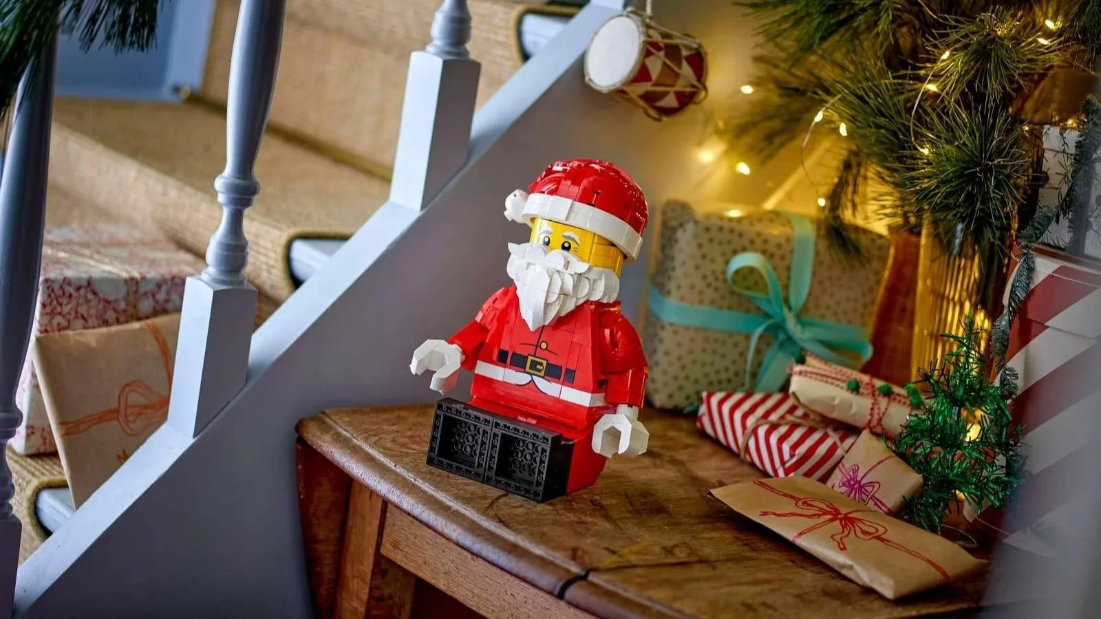 LEGO Up-Scaled Santa Minifigure sitting on a wooden table by a staircase, surrounded by wrapped gifts and warm tree lights