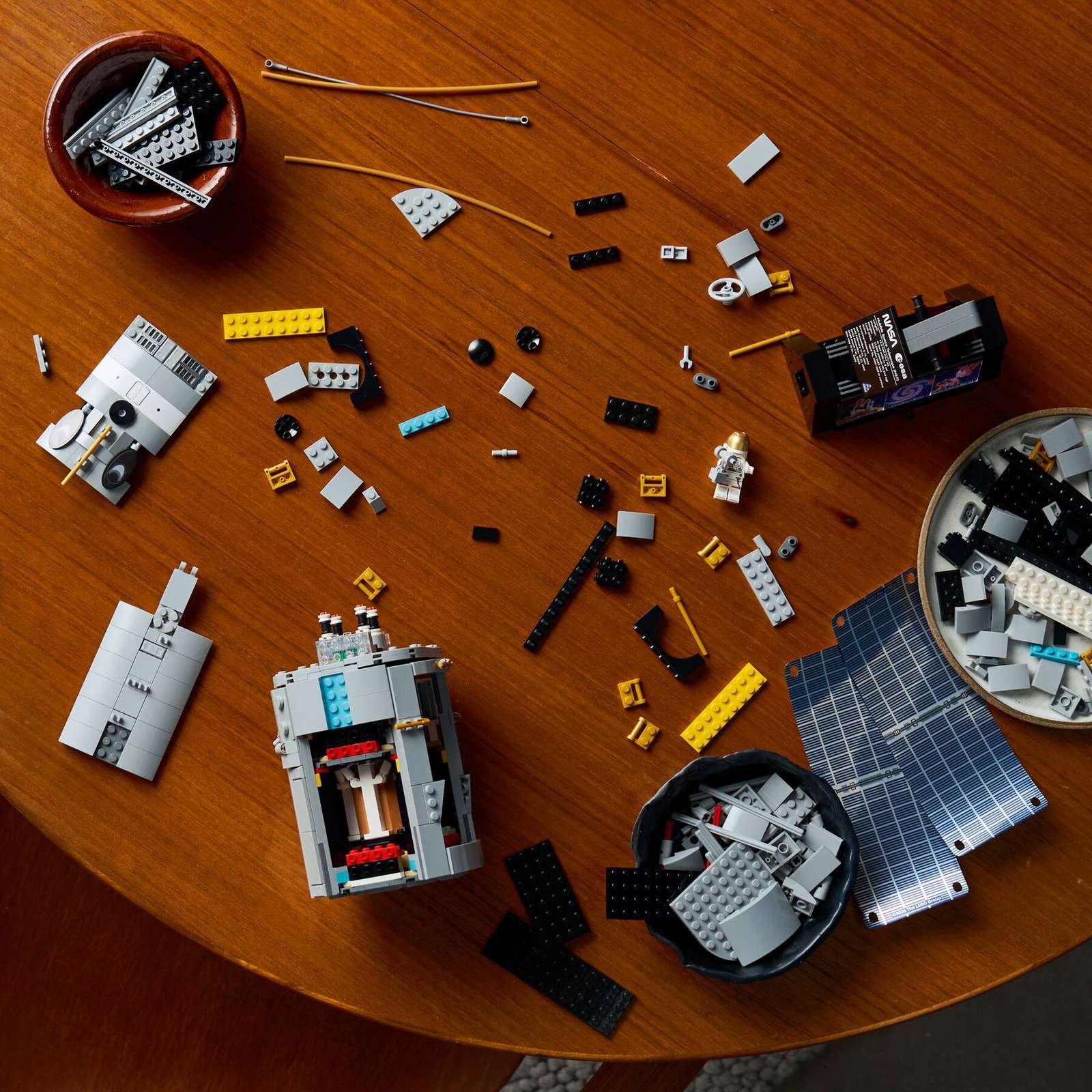 A LEGO Hubble Space Telescope build in progress on a round wood table. The gray telescope body and blue solar panels sit among sorted bricks, a NASA-branded module, and an astronaut minifigure wearing a gold helmet.