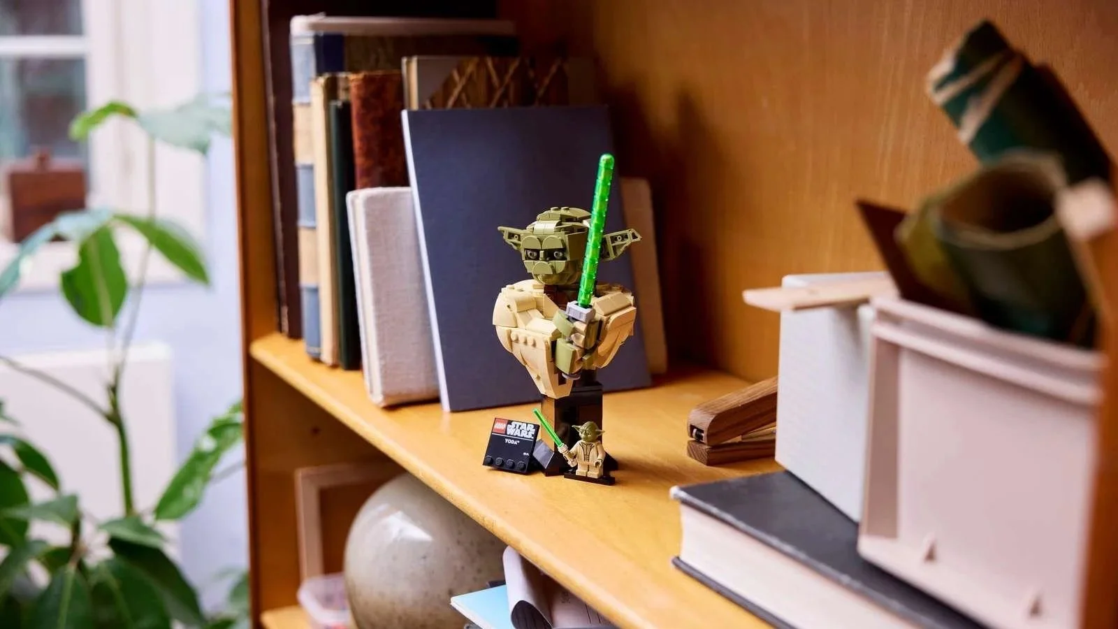 LEGO Yoda Bust holding a raised green lightsaber, displayed on a bookshelf next to its matching minifigure and nameplate