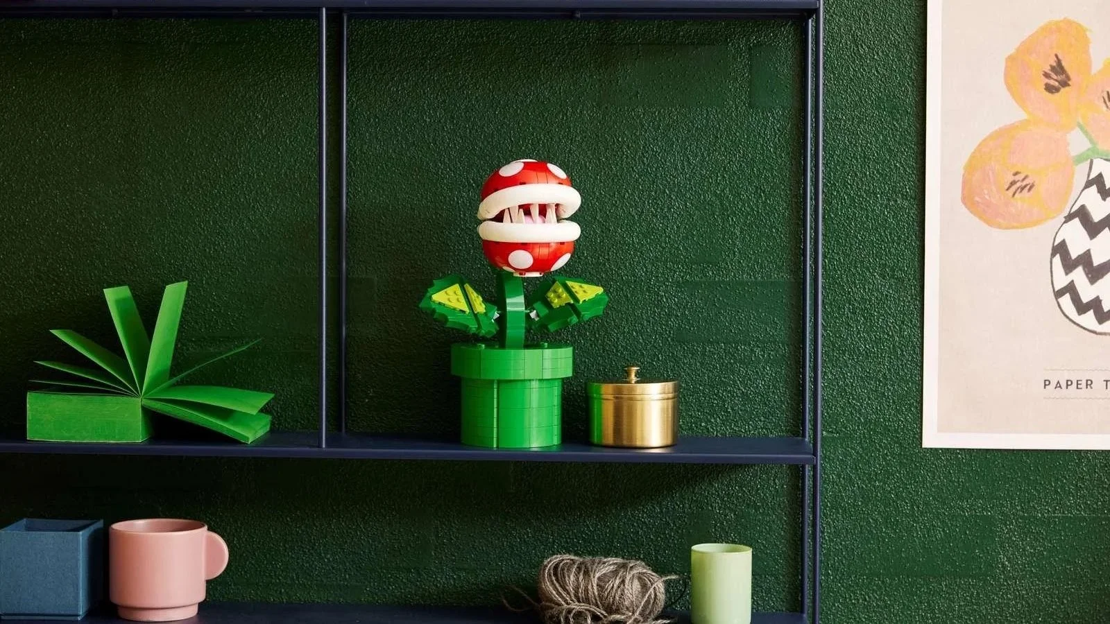 The LEGO Piranha Plant stands on a dark floating shelf against a green wall, framed by a folded paper sculpture and a botanical print