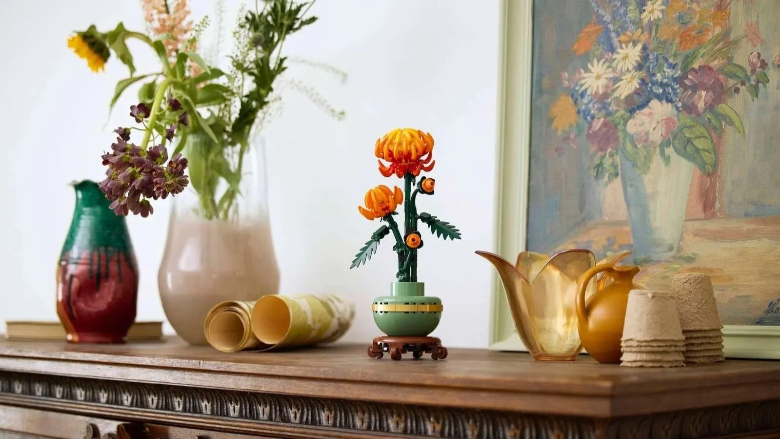 LEGO Chrysanthemum in its celadon pot on a wood sideboard, styled with fresh-cut flowers, a brass pitcher, and a framed floral painting