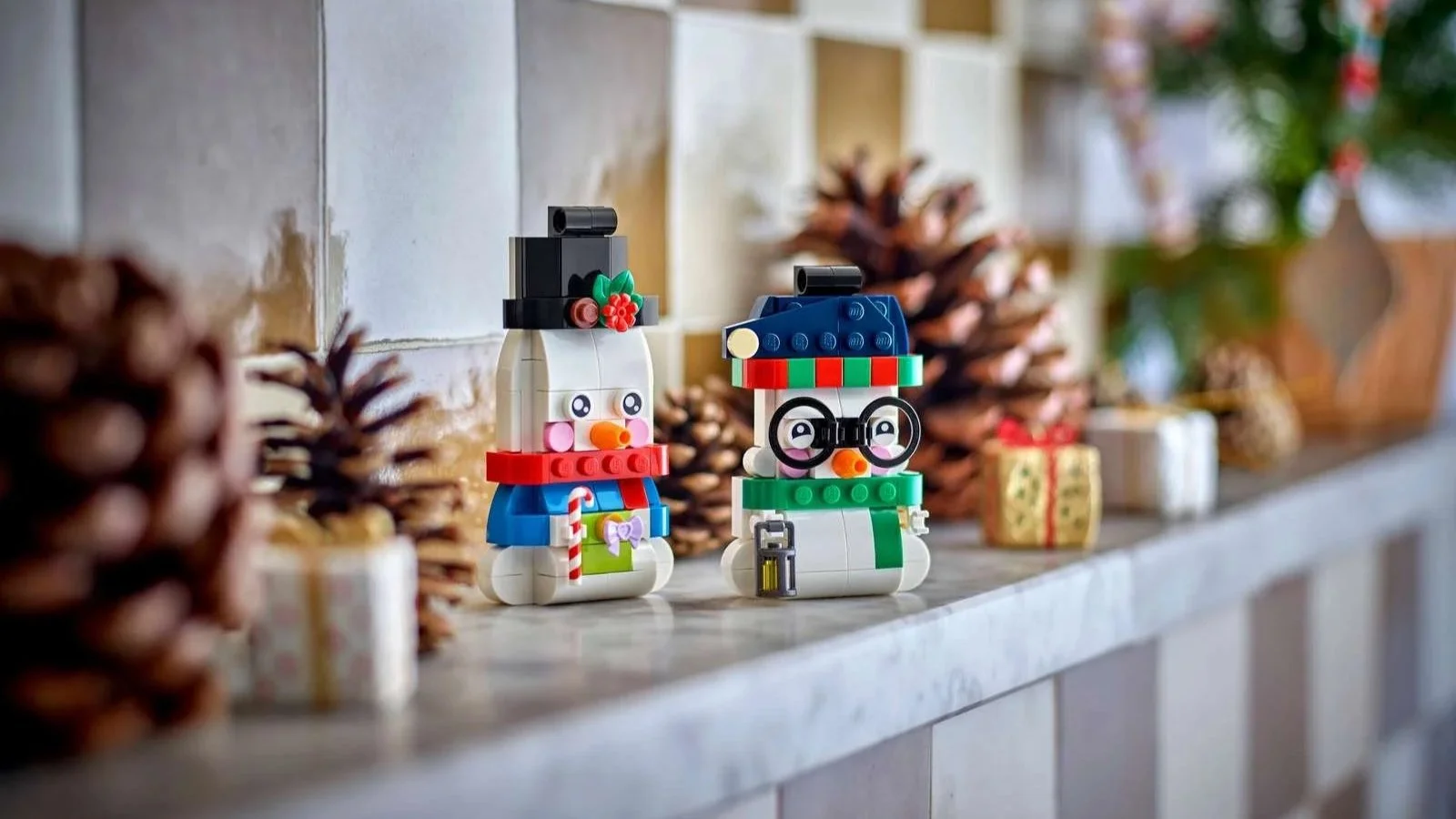 Two LEGO snowman ornaments, one in a top hat holding a candy cane, one in round glasses with a lantern, on a mantel with pinecones