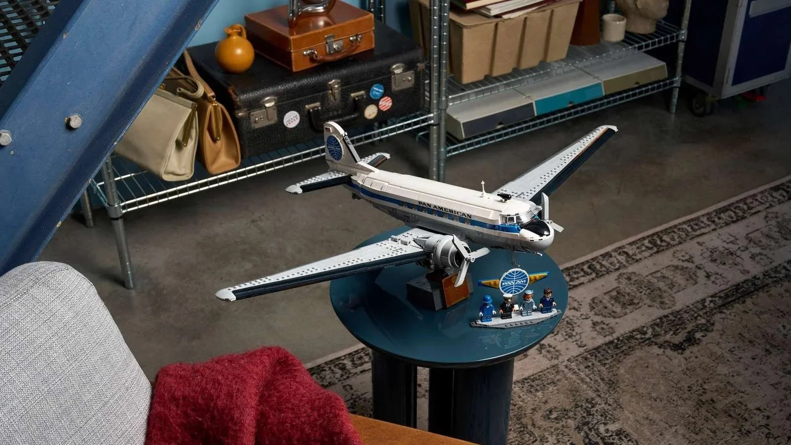 LEGO Douglas DC-3 Pan Am airliner model with flight crew minifigures, displayed on a side table with vintage luggage and a red throw blanket
