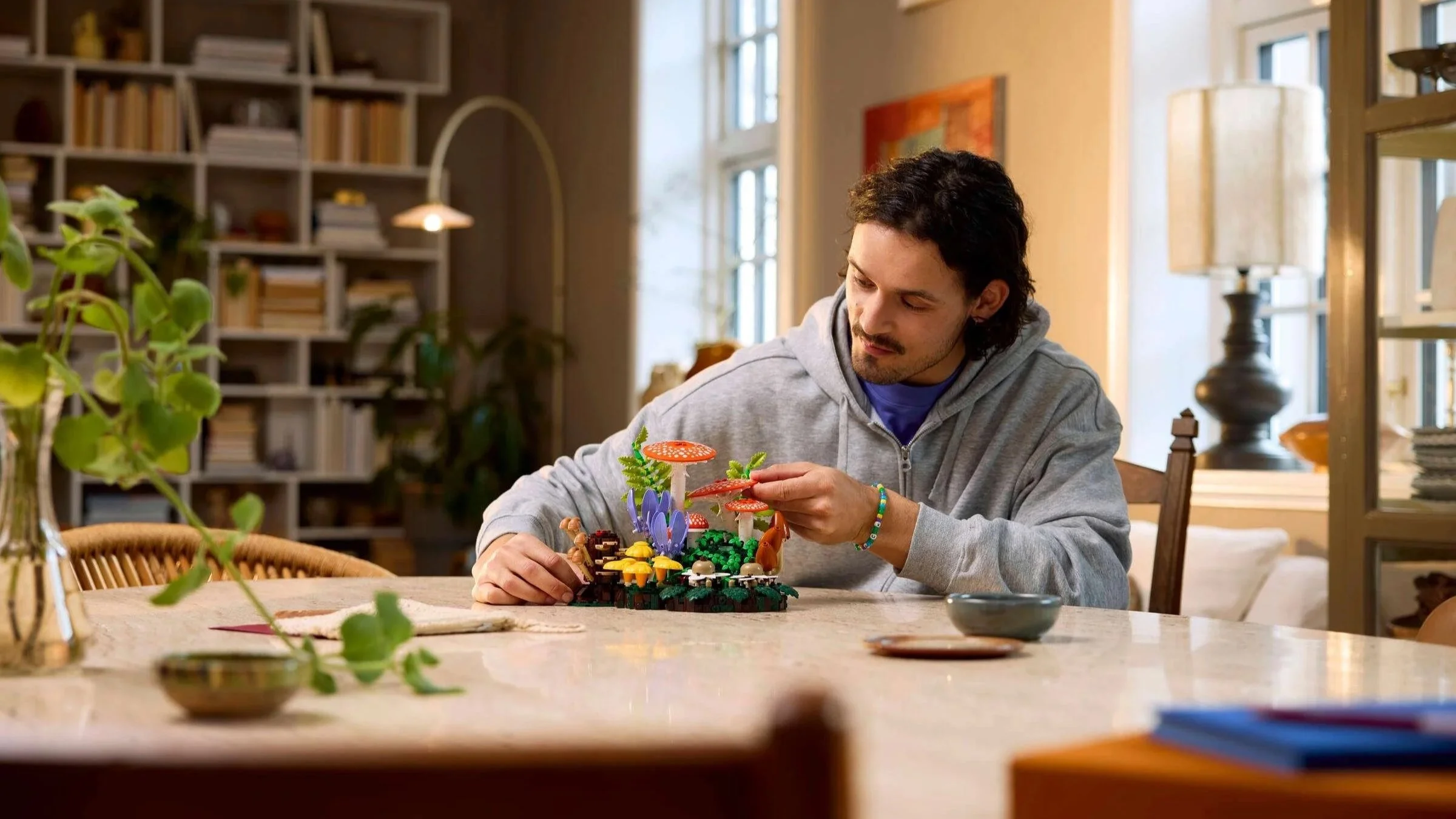 A man arranges a LEGO Woodland Mushrooms scene with red and white toadstools, ferns, and moss on a dining table