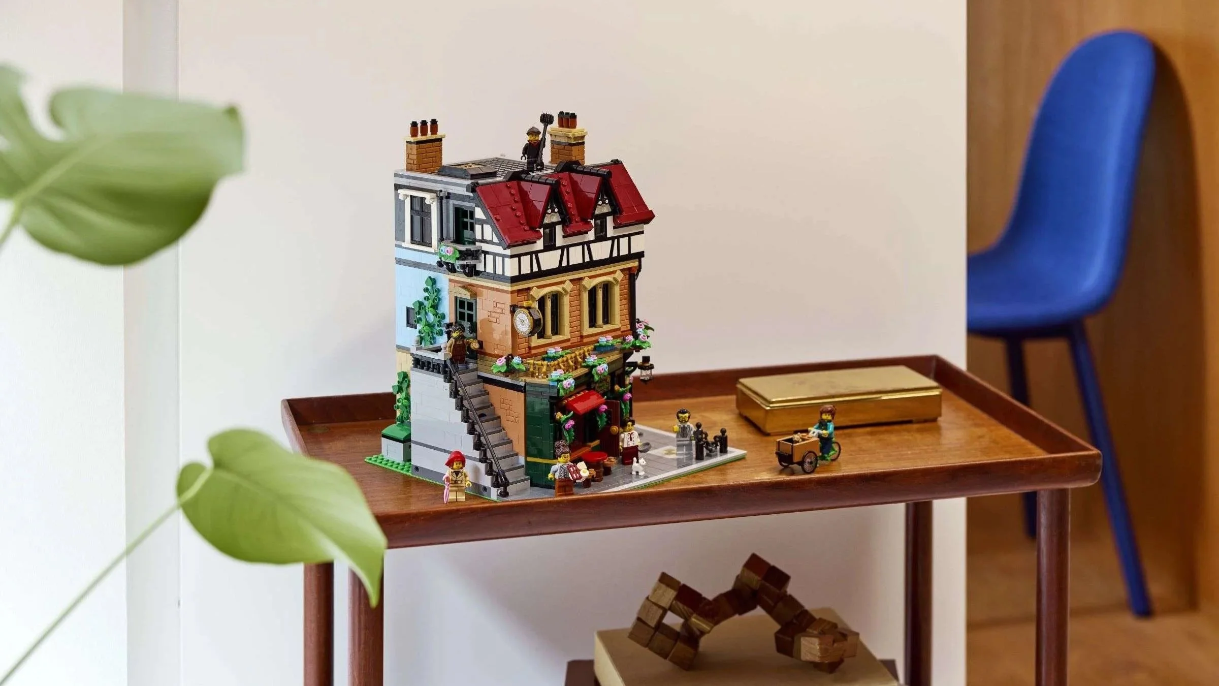 The LEGO Tudor Corner building from a side angle, showing its spiral staircase and rooftop chimney sweep figure, displayed on a wood cart beside a gold box